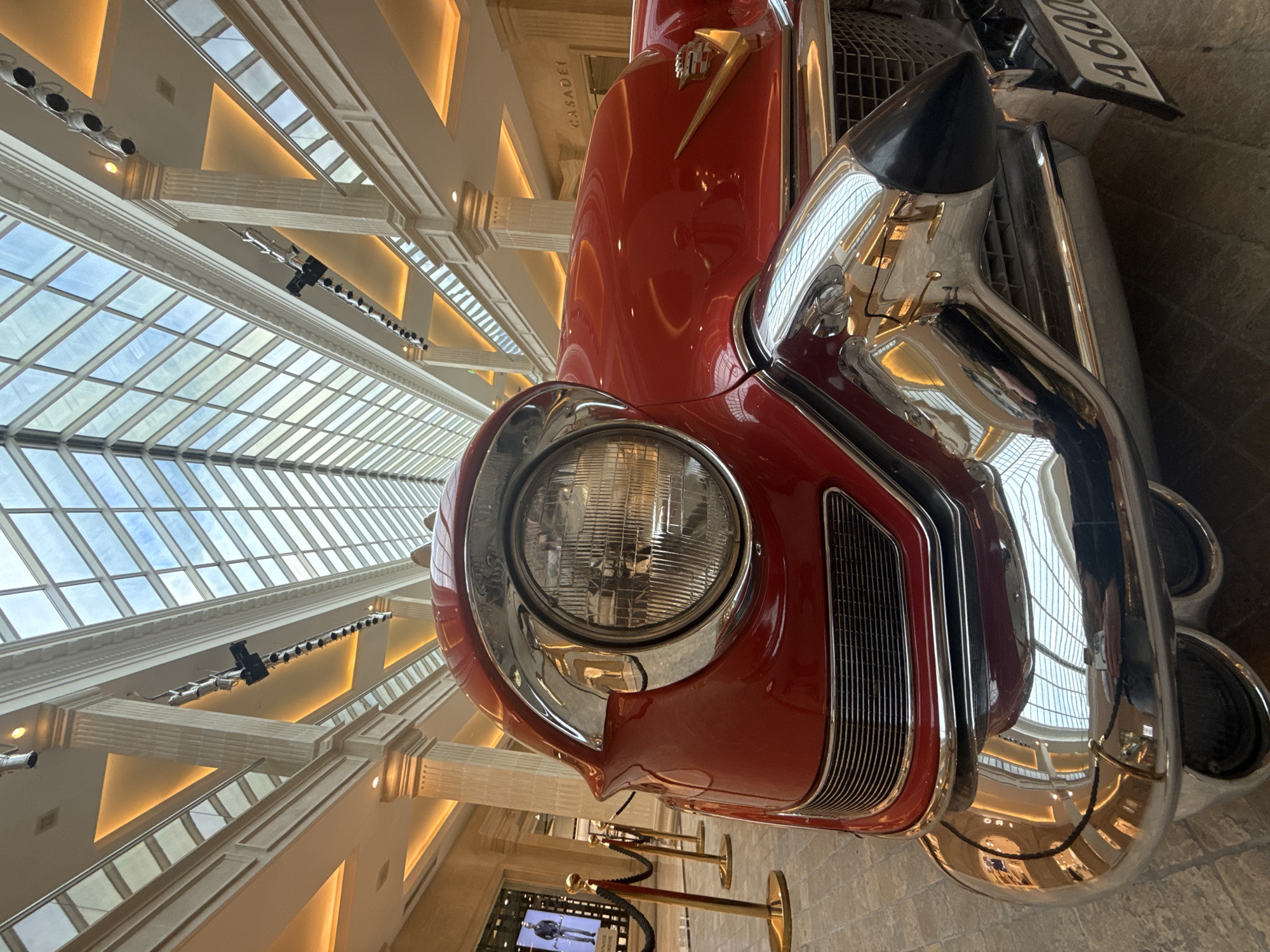 The image shows a close-up, low-angle view of the front of a vintage red car, possibly a Cadillac, displayed indoors. The car is the primary subject, with its polished chrome bumper, headlight, and grille prominently featured. The car's reflection is visible on the shiny surfaces, showing parts of the interior of the building.
The setting appears to be a grand, well-lit atrium or exhibition space, characterized by a high, vaulted ceiling made of glass panels, allowing natural light to filter in. Supporting columns with ornate detailing and illuminated ceiling features are visible, suggesting a luxurious architectural environment. The lighting is warm and diffused, indicative of indoor lighting, possibly supplemented by the natural light from the skylight.
There are no people actively engaged in activity in the frame. The scene is static, suggesting the car is on display. The overall mood is one of appreciation for classic automotive design and architectural grandeur.
Visible text includes "CASADEI" on the car's body and a license plate that reads "A600" followed by illegible characters. The context provided indicates the location is Khimki, Russia. The time of day cannot be determined definitively, but the indoor setting and lighting suggest it could be daytime. The weather is not discernible from the image.