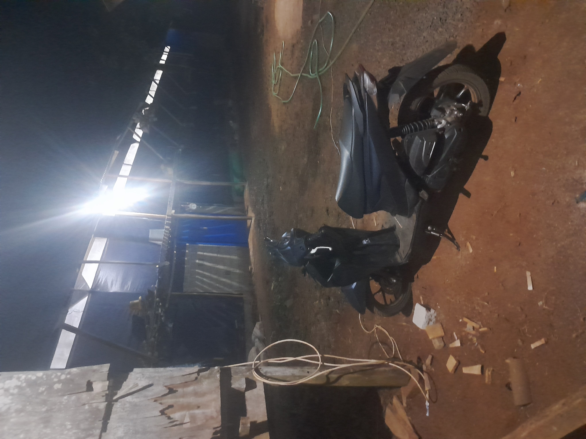 In Labuhan Ratu Lima, Indonesia, a nighttime scene unfolds in an outdoor, partially constructed or utilitarian area. A dark-colored scooter is parked on a dirt ground surface. Adjacent to the scooter, a coiled white wire is draped over a wooden post. Further to the right, a green hose lies coiled on the ground. The background features a structure with a corrugated metal roof, partially covered by blue tarpaulin. Several light panels are visible along the roofline, and a bright light source emanates from the upper left section of the structure, casting illumination on the scene. Scattered pieces of wood and a small white object are present on the ground near the scooter. The overall environment appears to be undeveloped or under construction, with visible wooden elements and rough terrain.