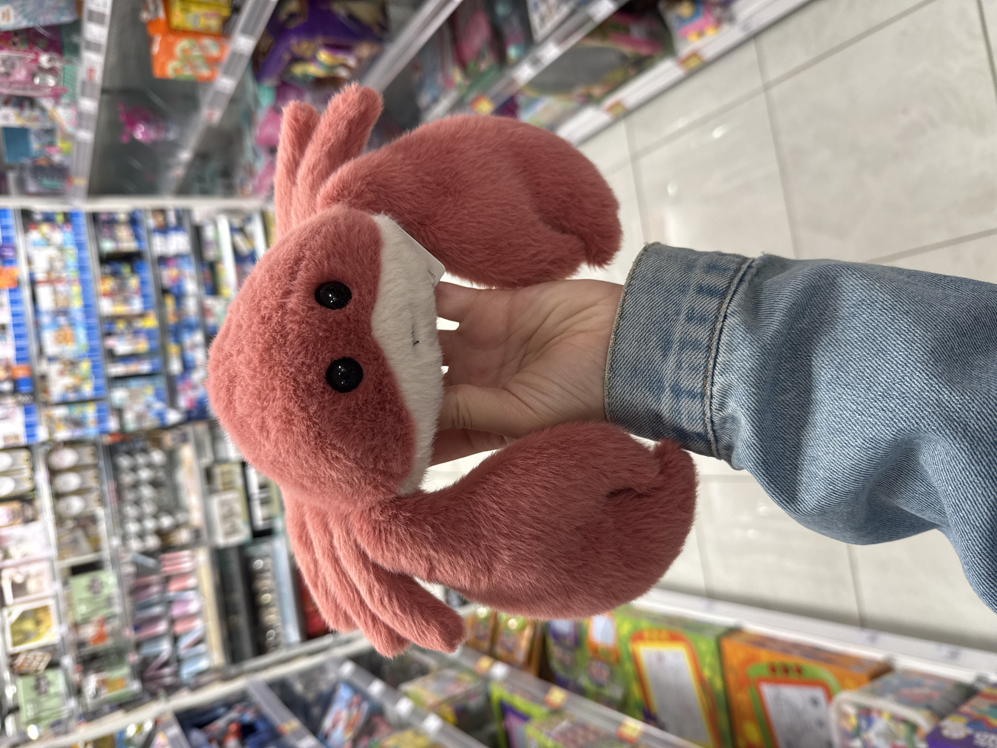 The image displays a close-up view of a person's hand holding a vibrant pink, plush, crab-shaped toy or mitten. The hand, wearing a light blue denim jacket sleeve, grasps the soft item, making its black button-like eyes and white belly visible. The plush crab has a distinctly fuzzy texture and several soft, rounded appendages resembling claws or legs.
The setting is an indoor retail environment, likely a supermarket or a toy store. The background features numerous blurred shelves stocked with various packaged products, suggesting a wide array of items. The flooring consists of light-colored, possibly white or grey, tiles. The overall lighting appears to be typical artificial indoor illumination. The activity depicted is a person holding or examining the plush item within this store. There is a small white tag visible on the plush, but no text on it or on the background products is discernible due to the shallow depth of field. The image was captured in the City of Zagreb, Croatia.