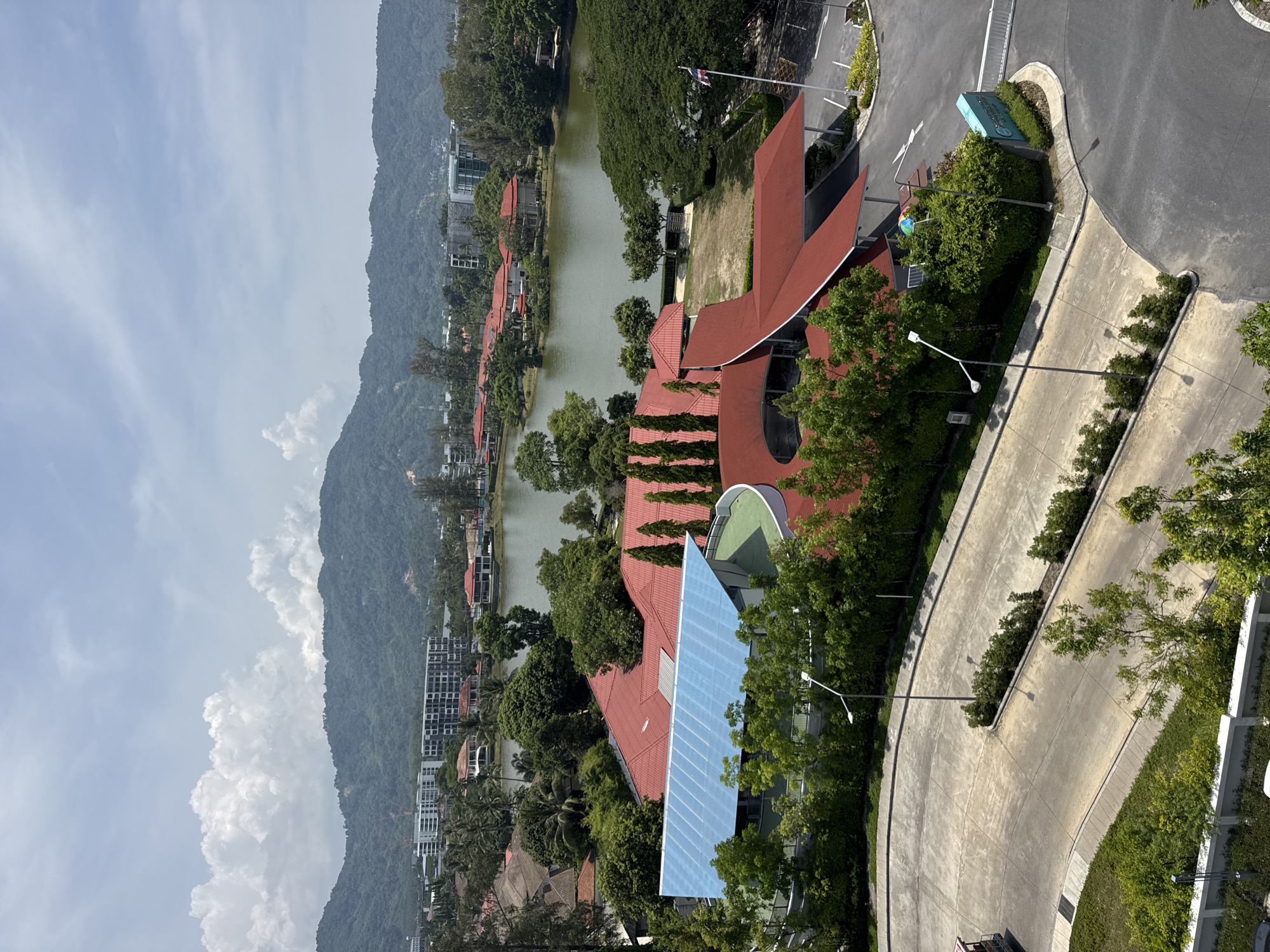 The image shows an aerial view of Choeng Thale, Thailand. The scene includes mountains on the left side, with building clusters at the base. A small lake is visible between the buildings and a building complex with red roofs. A road curves through the landscape, lined with streetlights and landscaping, to the right of the building complex.