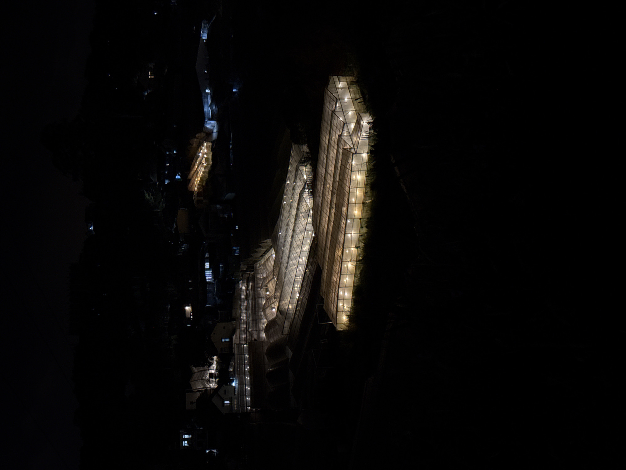The image shows a dark, aerial view of illuminated structures in what appears to be a city. Dominating the scene are three large, rectangular buildings or structures with grid-like patterns, brightly lit from within. The buildings are aligned diagonally across the frame, with the largest one on the right side. To the left of these structures, there are other, smaller illuminated buildings and what appears to be a road or pathway lined with lights. The rest of the image is mostly dark, indicating a night scene. Based on the location provided, the structures could be greenhouses or agricultural buildings in Lâm Viên - Đà Lạt, Vietnam.