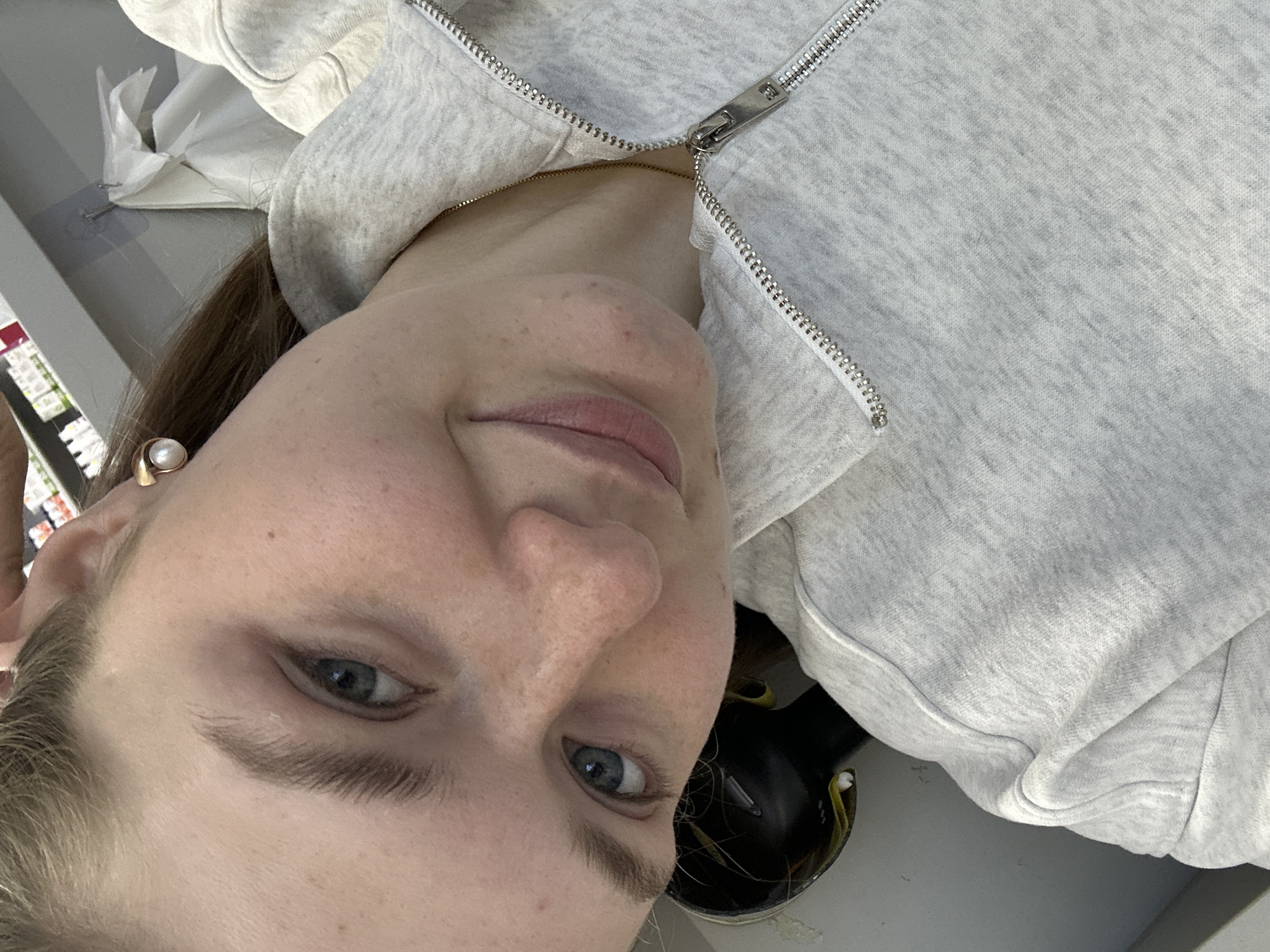 The image shows a selfie of a woman with light skin, blue eyes, and brown hair. She is wearing a light gray zip-up hoodie. A pearl earring is visible on her left ear. Part of a pair of black sunglasses is also visible. The location appears to be indoors, on a light gray surface, possibly a counter.