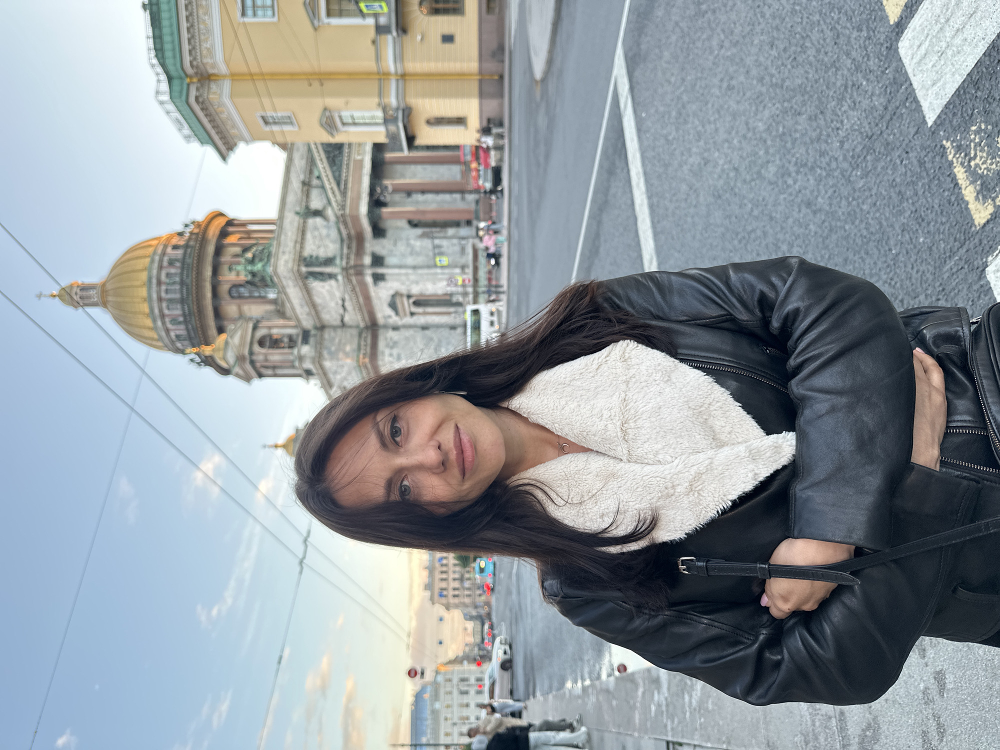 A medium shot captures a woman standing on a city sidewalk with her arms crossed, looking towards the camera with a slight smile. She has long, dark hair and is wearing a black leather jacket with a cream-colored fuzzy lining around the collar and lapels. A thin strap of a bag is visible across her chest.
Behind her, a wide street with traffic lights and some vehicles is visible. In the background, the iconic golden dome of Saint Isaac's Cathedral dominates the skyline, with a yellow building to its right. The sky above is a pale blue with wispy clouds, suggesting it is late afternoon or early evening. The street is paved with asphalt and marked with white and yellow pedestrian crossing lines. Several other people and buildings are present in the distance, indicating a bustling urban environment in Saint Petersburg, Russia.