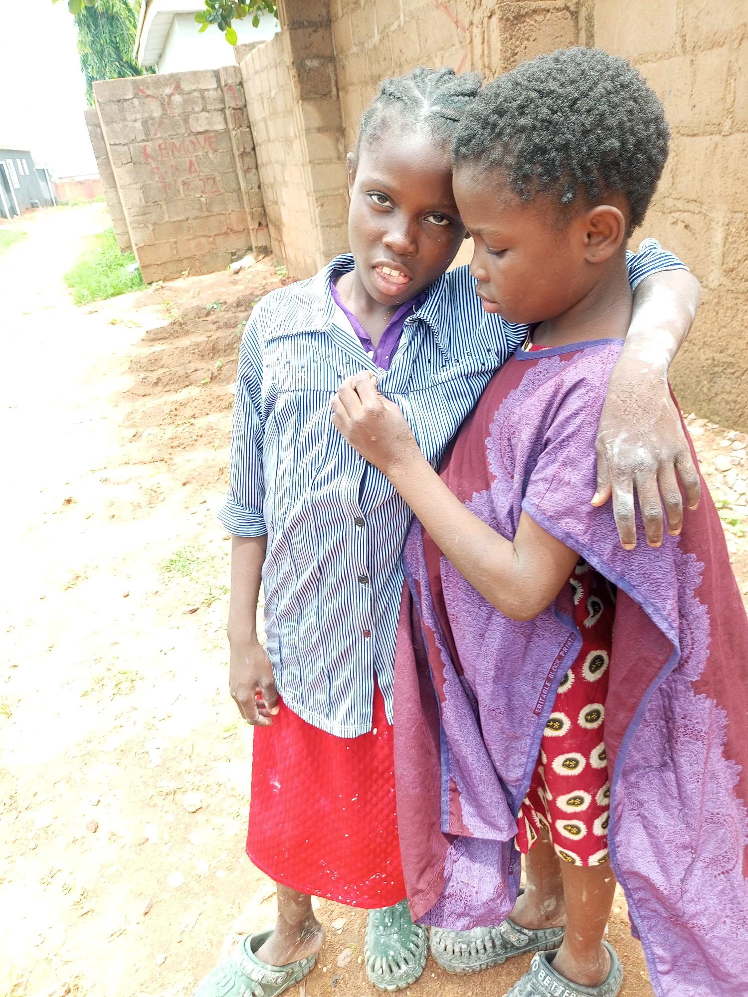 Two young Nigerian girls are standing outdoors in what appears to be a residential area in Tempure, Nigeria. The girl on the left, wearing a blue and white striped shirt and a red skirt, is looking towards the camera with her mouth slightly open. Her friend, on the right, is embracing her from behind, with her arm draped over the other girl's shoulders. She is wearing a purple garment over a red and white patterned dress. Both girls have dirt on their arms and legs, suggesting they have been playing outside. The background consists of a brick wall with some writing on it and a dirt path.