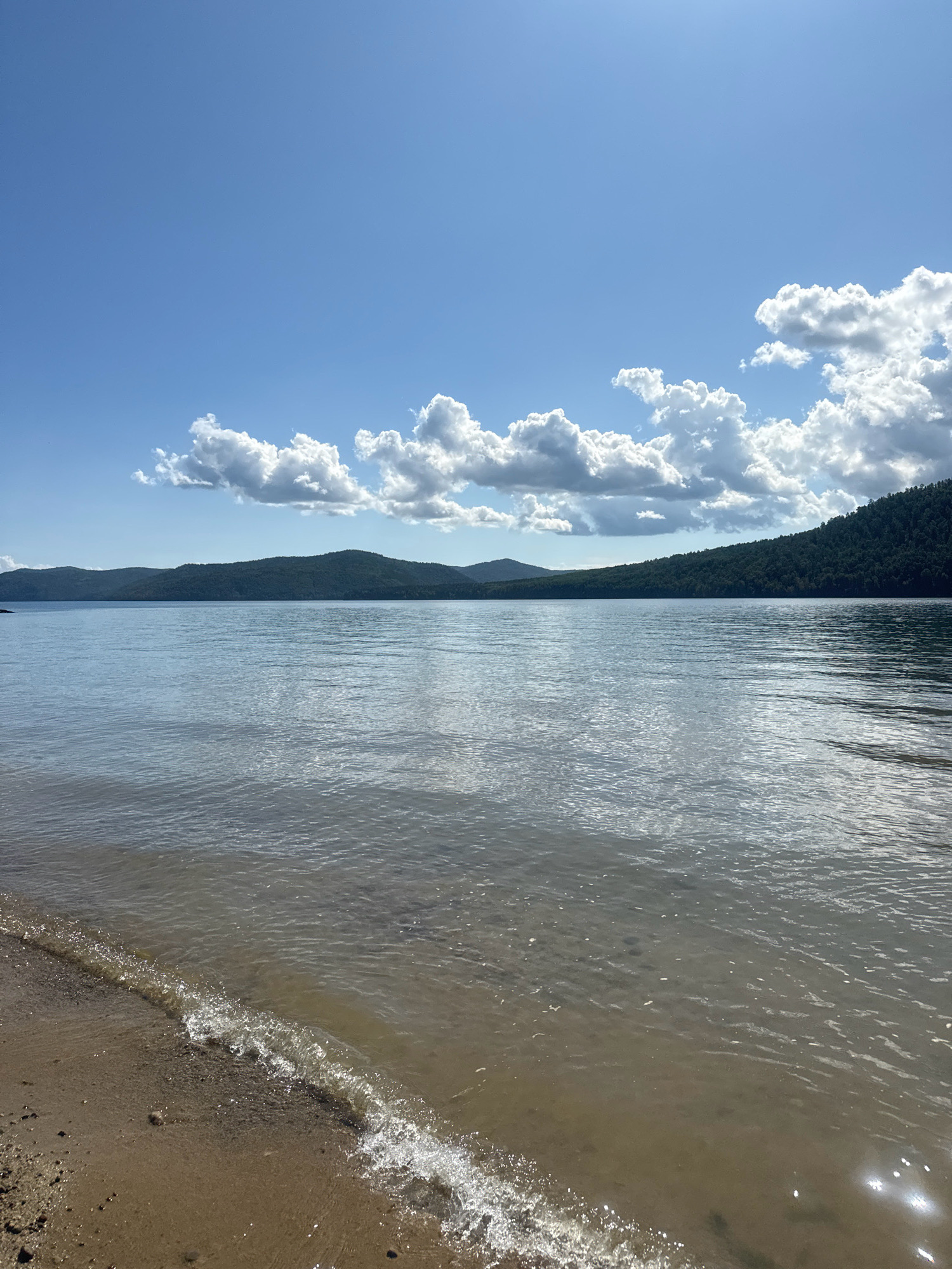 The image depicts a serene lake scene under a bright, partly cloudy sky. In the foreground, a sandy shoreline meets the calm, shallow water, with a small wave gently lapping at the edge. The water is clear enough to see the sandy bottom beneath. Across the expanse of the lake, a line of lush, green, forested hills forms the distant horizon. The sky is a vivid blue, punctuated by fluffy white cumulus clouds that cast shadows on the hills and reflect on the water's surface. The overall impression is one of natural beauty and tranquility. The location is identified as the Angara River near the "Taltsy" Museum in Irkutsk, Russia.