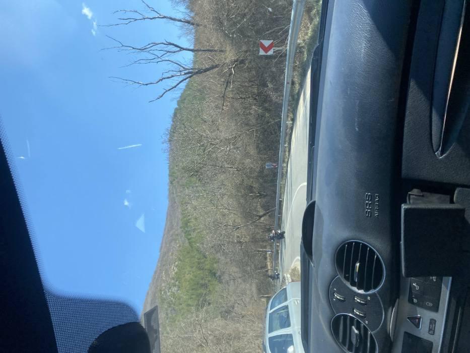 The image captures a scene from inside a vehicle, looking out through the windshield onto a winding mountain road in Konyavo, Bulgaria, on a clear, sunny day. The foreground shows the vehicle's dark gray dashboard, featuring circular air vents on the left, control buttons, an "SRS AIRBAG" label on the passenger side, and a black mounting device on the lower right. Reflections are present on the windshield.
The asphalt road curves to the right, with a metal guardrail lining its right edge. Behind the guardrail, a red and white chevron sign indicates the sharp right turn. Three individuals on motorcycles are visible on the road ahead, navigating the curve. The most prominent motorcyclist is in the center, wearing dark gear, with two other motorcyclists further behind. A silver-colored station wagon is partially visible on the left side of the road, appearing to be stationary or moving slowly.
The background is dominated by a range of hills or mountains covered predominantly with bare deciduous trees, suggesting late winter or early spring. Patches of green evergreen trees are visible on the upper slopes to the left. The sky is clear and blue, with a faint white contrail from an aircraft visible in the upper right quadrant. Several prominent bare trees with distinct upward-reaching branches are positioned on the right side of the scene, behind the guardrail.