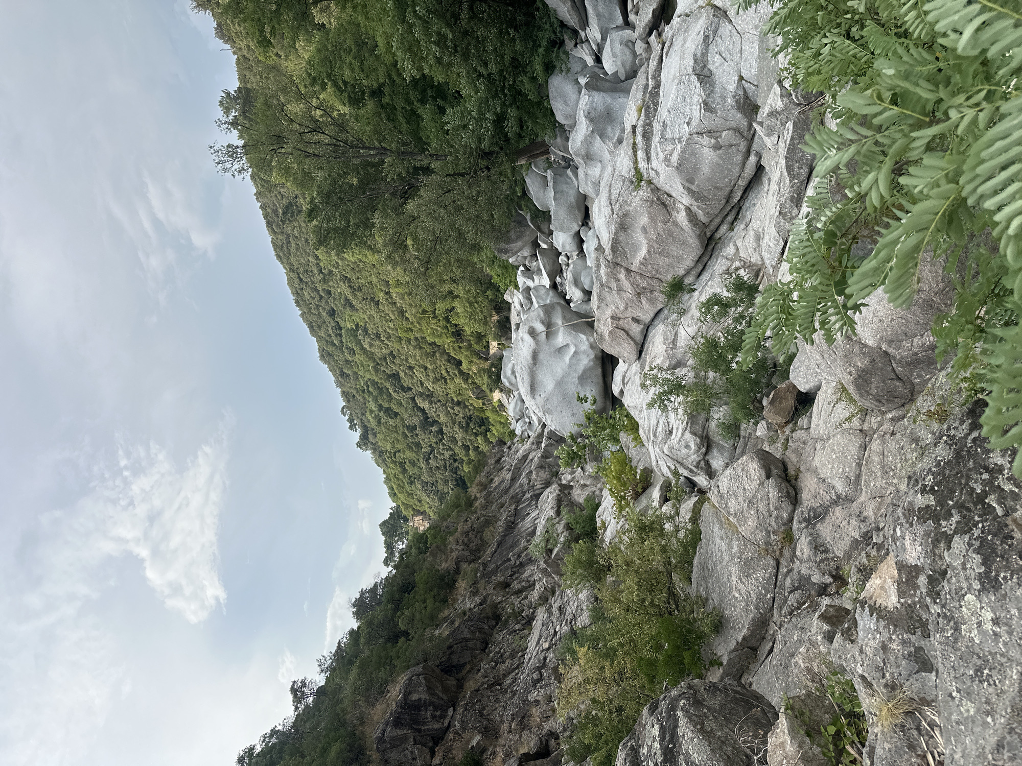 This media file depicts a rocky, mountainous landscape covered in dense green foliage. The scene is dominated by large, weathered grey rocks, some appearing smooth and rounded, others jagged and rough. The vegetation is lush and varied, with trees and smaller plants growing among the rocks and covering the steep hillsides. The perspective is from a high vantage point, looking down into a ravine or gorge. In the distance, nestled among the trees on a hillside, a small, light-colored building with a red roof is visible.
The time of day appears to be daytime, with a bright, overcast sky and scattered white clouds. The lighting suggests it might be late morning or early afternoon. The weather seems to be mild, with no strong indications of rain or extreme temperatures. The overall impression is one of a wild and natural environment, possibly a national park or a remote natural area. The rugged terrain and the presence of the building suggest a location that is accessible but still retains a sense of wilderness. There is no visible text in the image.