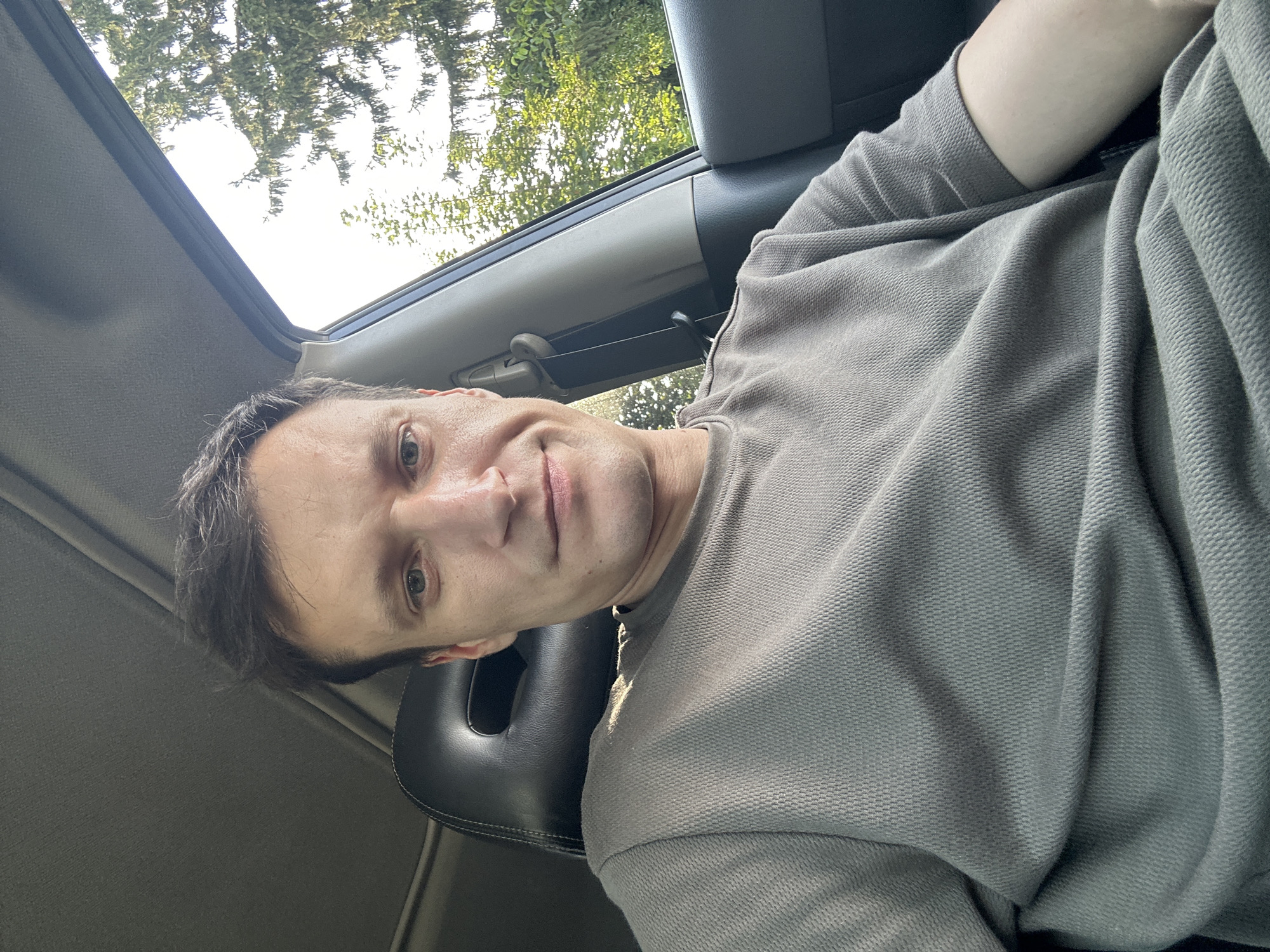 A young man is sitting in the driver's seat of a car, looking directly at the camera. He has short, dark brown hair and light-colored eyes. He is wearing a plain, grey, short-sleeved t-shirt with a textured knit. The interior of the car is visible, including a dark headrest and part of the dashboard. Through the open car window, a bright, overcast sky and green trees are visible, suggesting an outdoor setting, possibly in Shatava, Ukraine, where deciduous trees are common. The man's expression is neutral, with a slight, almost imperceptible smile. The lighting suggests it is daytime, with sunlight filtering in from the side. The image appears to be a selfie.