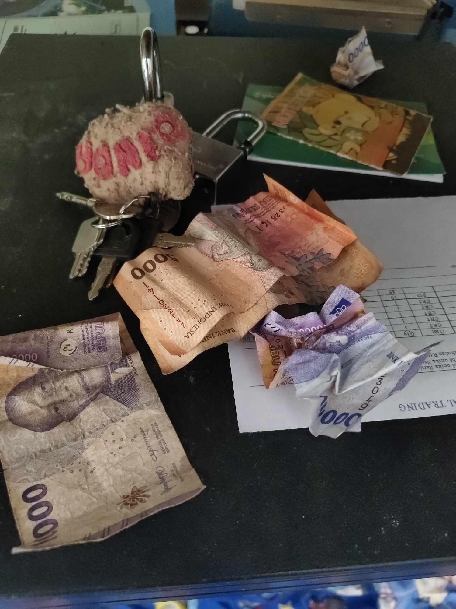 A close-up, high-angle shot shows a dark, textured surface, likely a table, in an indoor setting. Scattered across the surface are several Indonesian Rupiah banknotes in various denominations and conditions. Some are folded, while others are crumpled. Among the currency, a set of keys with a fluffy, pink-lettered keychain is visible. Also present are two padlocks, one of which is secured to the keychain. A small, folded piece of paper with the numbers "0000" printed on it is also on the surface, as is a small, illustrated card that appears to be a sticker or part of a larger item. A white sheet of paper with a grid of columns and rows is partially visible in the foreground, with text that appears to be in English, including "TRADING." The overall impression is a casual arrangement of personal items, possibly in a home or office environment.