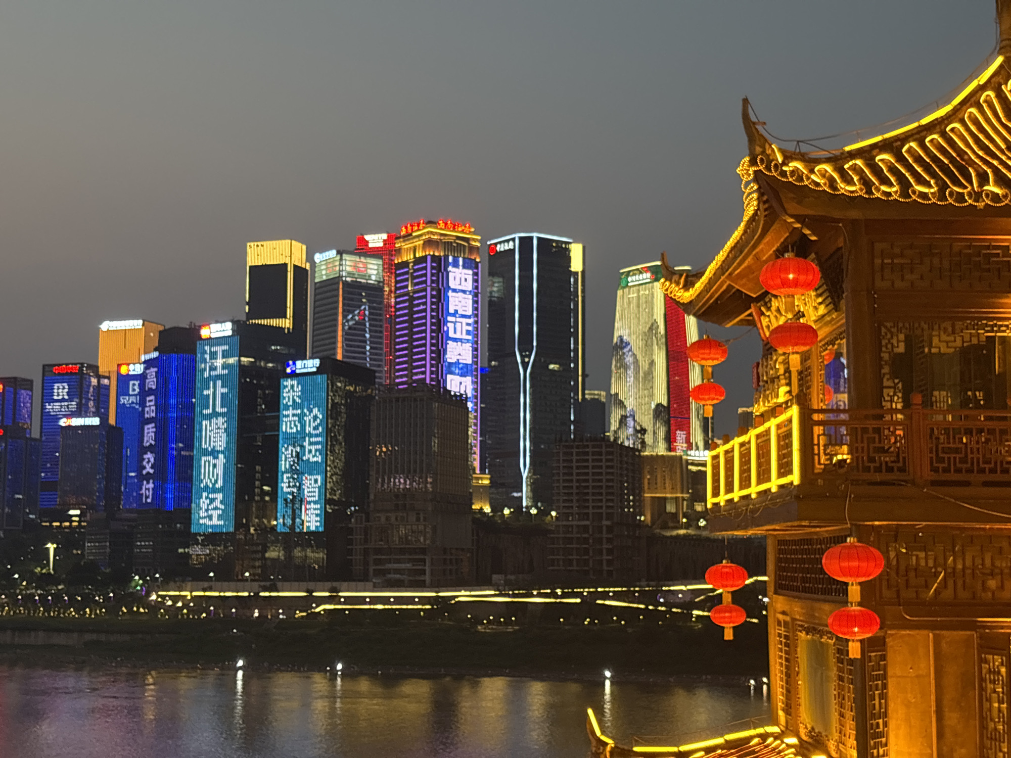 The photograph shows a nighttime view of a cityscape, likely in the Yuzhong District of Chongqing, China, as evidenced by the architectural style and visible signage in Chinese characters. Dominating the scene are several skyscrapers illuminated with colorful lights and digital displays, some of which show Chinese characters. In the foreground, a traditional Chinese-style pavilion or structure is adorned with glowing yellow lights and red lanterns, providing a cultural contrast to the modern skyline. The water reflects the city lights, adding to the overall brightness of the scene. The sky is dark. The image is captured from a low angle, emphasizing the height of the buildings and the scale of the city. The photograph composition is a combination of modern and traditional elements.