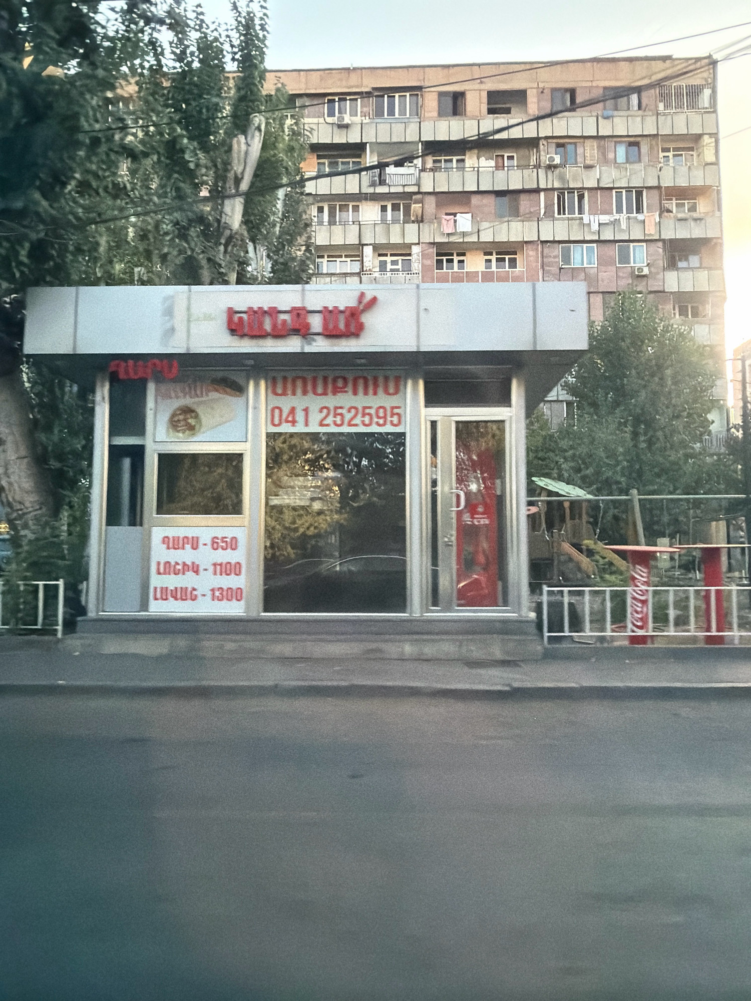 The image captures a street-level view of a small food kiosk in Yerevan, Armenia, situated in front of a multi-story apartment building. The kiosk has a modern, angular design with a white metal exterior and large glass windows and doors. Red lettering on the facade reads "ՇԱՎԱՐՏ" (Shavart), indicating the name of the establishment, and below it, a phone number "041 252595". A menu with prices in Armenian script is displayed on the left side of the kiosk, listing "ՇԱՎԱՐՏ - 650", "ԼՈՆԵԲ - 1100", and "ԼԱՎԱՇ - 1300". Inside the kiosk, a picture of a shawarma is visible. A red Coca-Cola refrigerator is positioned next to the entrance door.
To the right of the kiosk, a white metal fence encloses a small outdoor seating area with a red table and chairs, featuring Coca-Cola branding. Behind this area, a children's play structure is visible. The background is dominated by a tall, dated apartment building with numerous balconies and windows, typical of Soviet-era architecture found in Yerevan. Trees with dense foliage partially obscure the building. The street in the foreground is asphalt, and the sky above is a pale, hazy blue. The overall scene suggests a typical urban environment in Yerevan.