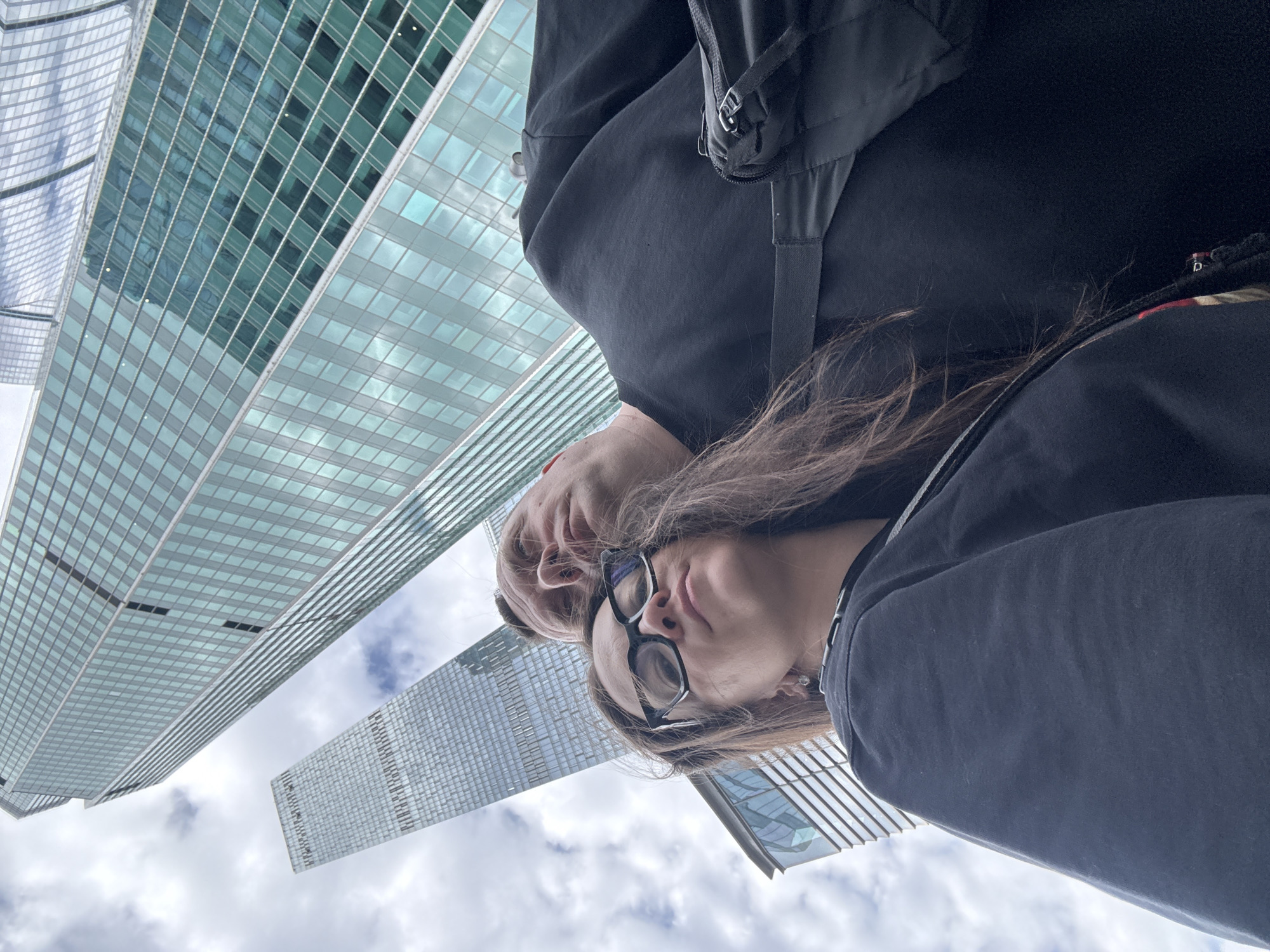 Two individuals stand closely together, looking upwards at modern skyscrapers. The taller building features a grid-like pattern of glass windows, reflecting the cloudy sky. The lower-angled buildings also have similar architectural styles. The individual in the foreground wears glasses and a dark-colored jacket. The second person is partially obscured.