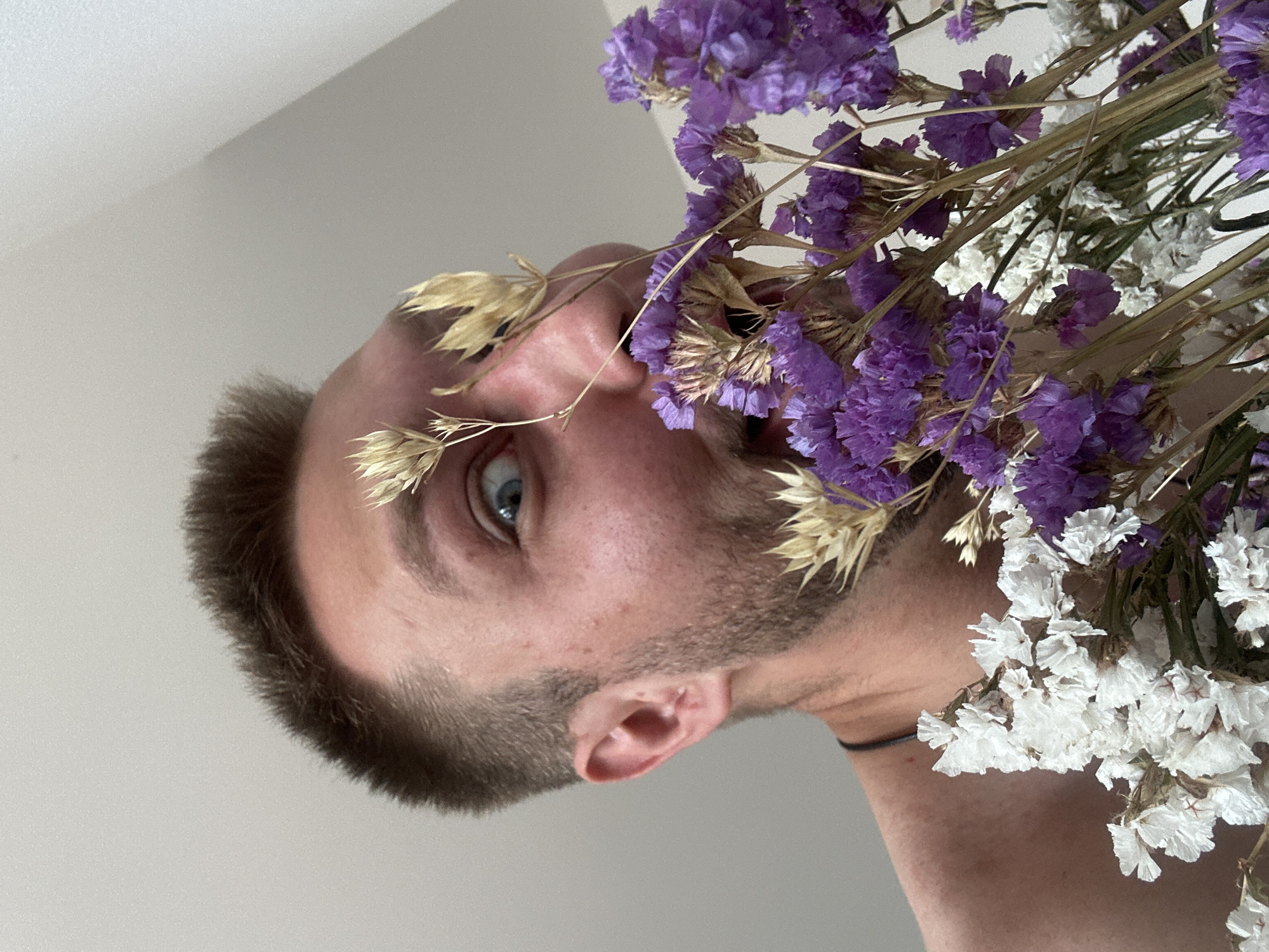 A close-up, low-angle shot shows a man with short brown hair and blue eyes holding a bouquet of dried flowers, predominantly purple and white, in front of his face. He appears to be in a bedroom in Zvezdara Urban Municipality, Serbia, as indicated by the plain, light-colored wall and ceiling, suggesting an indoor residential setting. The man's mouth is slightly open, and his gaze is directed towards the camera with a playful or surprised expression. The dry flowers, some with dried grass stalks, obscure parts of his face. His bare shoulders are visible, and he is wearing a dark necklace. The lighting is soft and natural, coming from the side, creating subtle shadows. The focus is sharp on the man's face and the flowers, with the background slightly blurred.