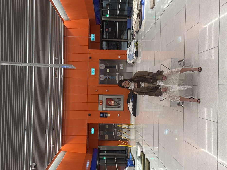 The image depicts a female individual seated centrally in an underground Tel Aviv light rail station, likely serving as a temporary shelter. The environment features bright orange paneled walls, a grey slatted ceiling with linear LED lighting, and light grey tiled flooring. Multiple sleeping mats, blankets, and personal belongings are scattered across the floor in the background and foreground, indicating an occupancy by multiple individuals not visible in the frame.
The central figure, a female with long dark hair, is seated upright on a light grey stone bench, facing the camera with a neutral expression. She wears a white ruffled high collar, an oversized brown jacket draped loosely over a lighter top, a long white semi-sheer skirt or dress, and black high-heeled sandals. Her hands are clasped in her lap.
In the background, visible features include two green "EXIT" (יציאה) signs above dark doors on either side of a central red fire extinguisher cabinet (כיבוי אש). A yellow collapsible barrier is present on the far left. A blue rectangular sign hangs from the ceiling. A CCTV camera is mounted on the ceiling in the upper left corner. The presence of Hebrew text on the exit signs and fire extinguisher cabinet confirms the location in Tel-Aviv, Israel.