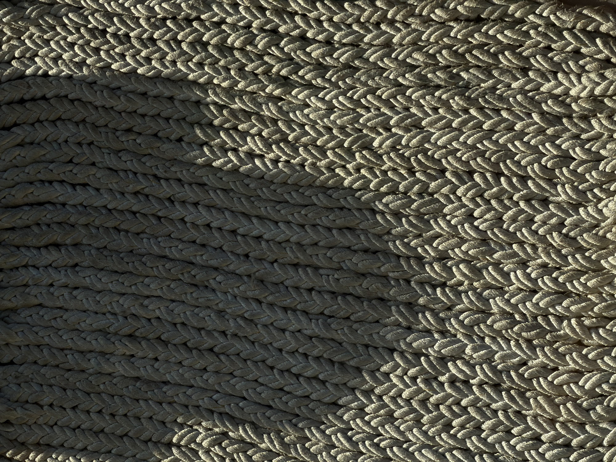 This is a close-up image of a tightly wound, thick rope. The rope appears to be beige or light gray in color. The texture of the rope is prominent due to the close proximity of the shot. The lighting is uneven, with a significant shadow covering approximately half of the frame. The shadow falls diagonally across the rope, creating a distinct contrast between the illuminated and shaded areas. The background is obscured by the rope, making it difficult to determine the setting or context of the image. There is no evidence of people or other objects in the picture. There is no identifiable information to determine the location in the city of Unknown, Unknown.