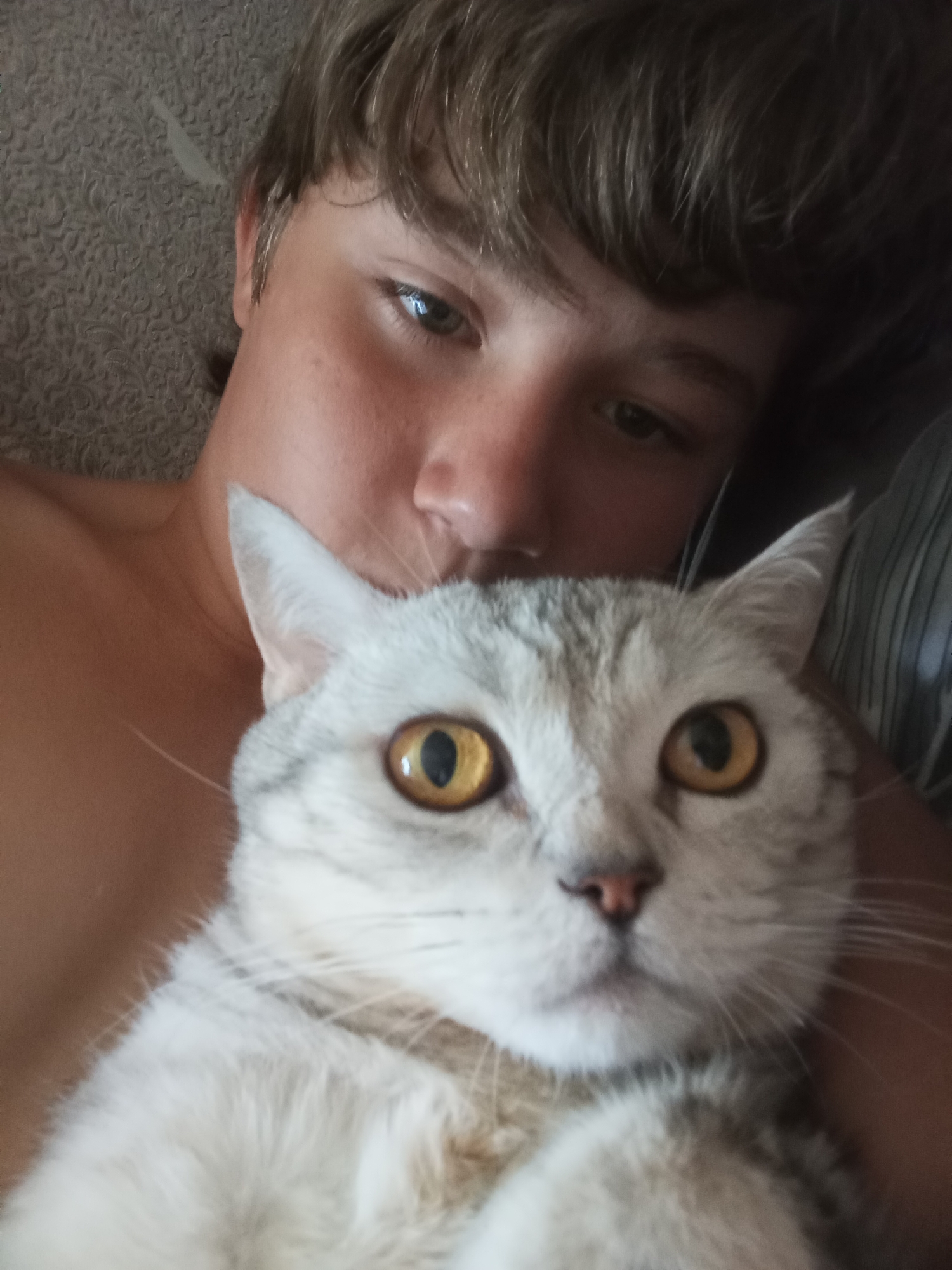 This is a close-up selfie taken by a young person with short, light brown hair, looking at the camera. The person is lying down, with their bare shoulders visible. They are holding a light-colored cat with distinctive yellow eyes and grey markings. The cat's face is prominently featured in the foreground. The background consists of a textured surface that appears to be a blanket or wallpaper, and a hint of striped fabric. The setting is likely indoors, possibly a bedroom. There are no discernible landmarks indicating a specific location in Sozonivka, Ukraine.