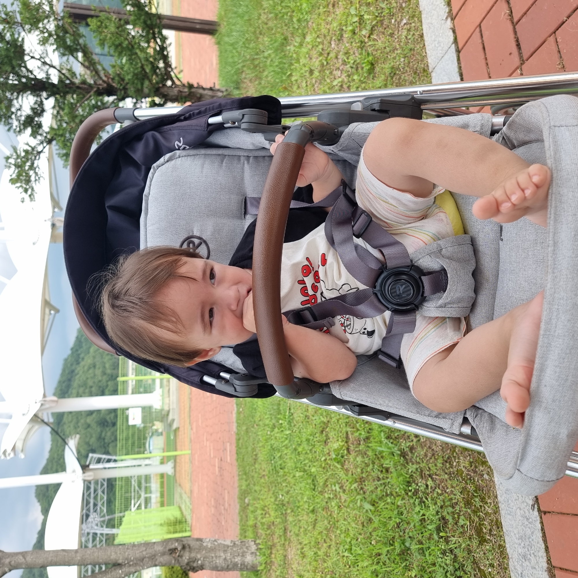 The image captures a close-up view of a toddler sitting in a stroller, outdoors in Dunnae-myeon, South Korea. The toddler, with brown hair, is looking down and biting their hand, wearing a black t-shirt and light-colored shorts with horizontal stripes. The stroller is gray with a brown handlebar. In the background, there is green grass, a brick pathway, trees, and a cloudy sky with some modern white structures that suggest a park or recreational area. The overall setting appears to be a public space.