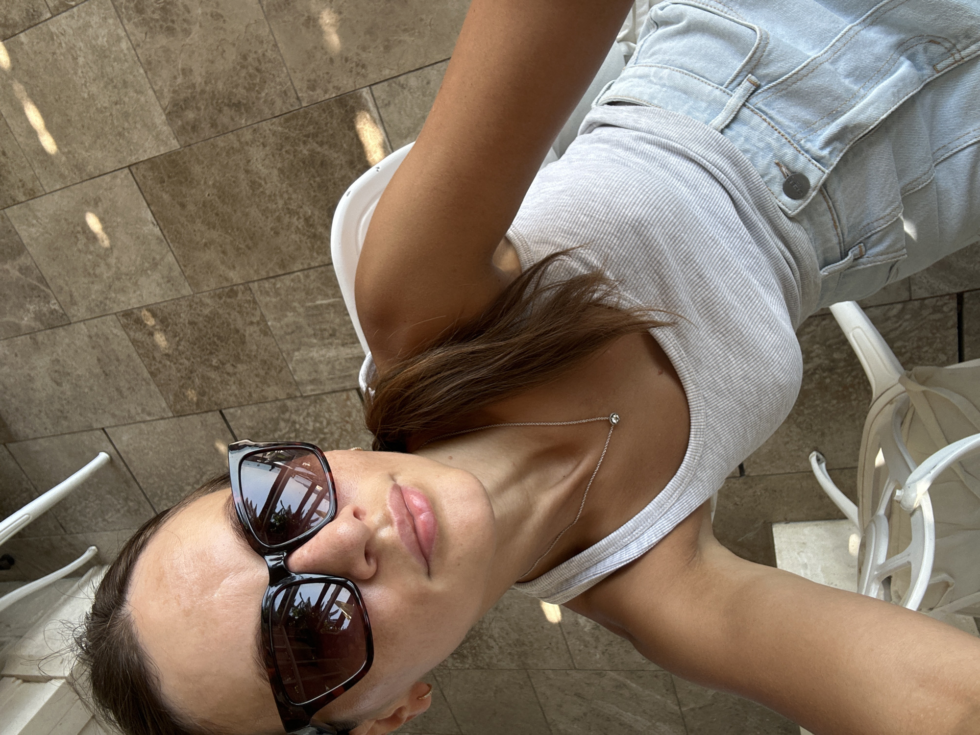 In Bečići, Montenegro, a woman is captured in a selfie, seated outdoors. She is wearing a light grey ribbed tank top and light blue denim shorts. Large, dark-framed sunglasses partially obscure her face, which is tilted slightly upwards. A delicate silver necklace with a single pendant rests on her collarbone. Her brown hair is pulled back into a low ponytail. She is sitting on a white chair, with the tiled floor of what appears to be an outdoor seating area visible beneath her. White chairs and table legs are partially visible in the background, suggesting a café or restaurant setting. Sunlight casts dappled shadows on the floor. The reflection in her sunglasses shows palm trees and a body of water, consistent with a coastal location.