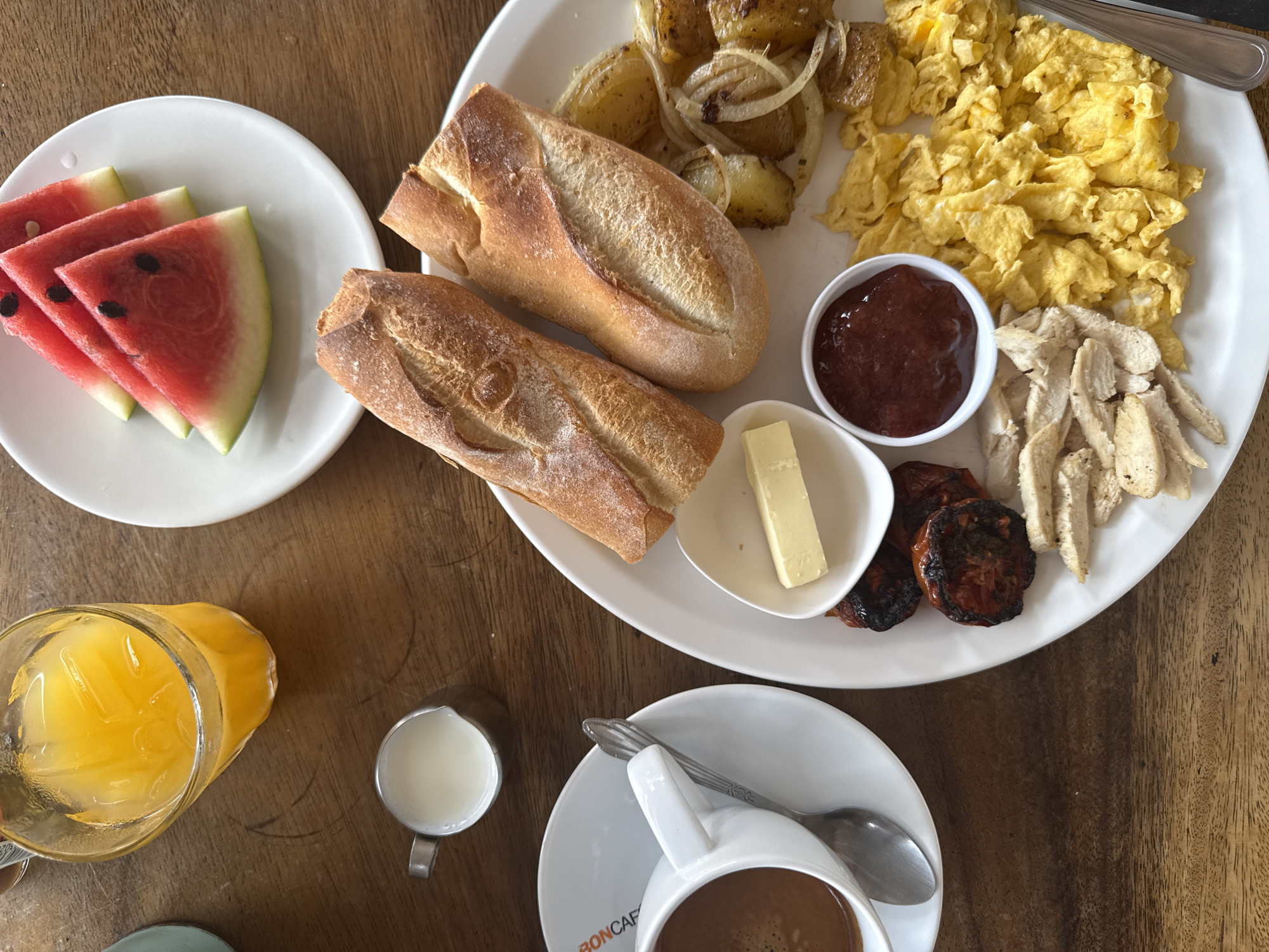 A high-angle, close-up shot shows a breakfast spread on a wooden table. In the upper left, a glass of orange juice filled with ice sits on the table. Next to it, a small stainless steel pitcher contains milk. A white saucer with a coffee cup and spoon is to the left of the pitcher. The coffee in the cup is dark brown. In the upper right, a white plate holds slices of watermelon. The main portion of the breakfast is on a large white plate in the center and right of the frame. This plate contains two French bread loaves, scrambled eggs, shredded chicken, roasted tomatoes, a small dish of jam, a small dish of butter, and sliced potatoes with onions. The scene is located in Baan Hua Thanon, Thailand. There are no people visible in the image.