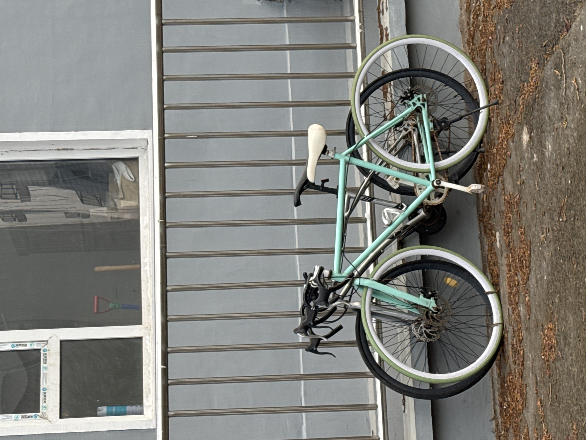 The image depicts a light-blue bicycle leaning against a gray building exterior in Zhongtun, China. The bike has white wheels and a white seat. A window is visible to the left, reflecting objects inside. Debris is present on the adjacent concrete surface.