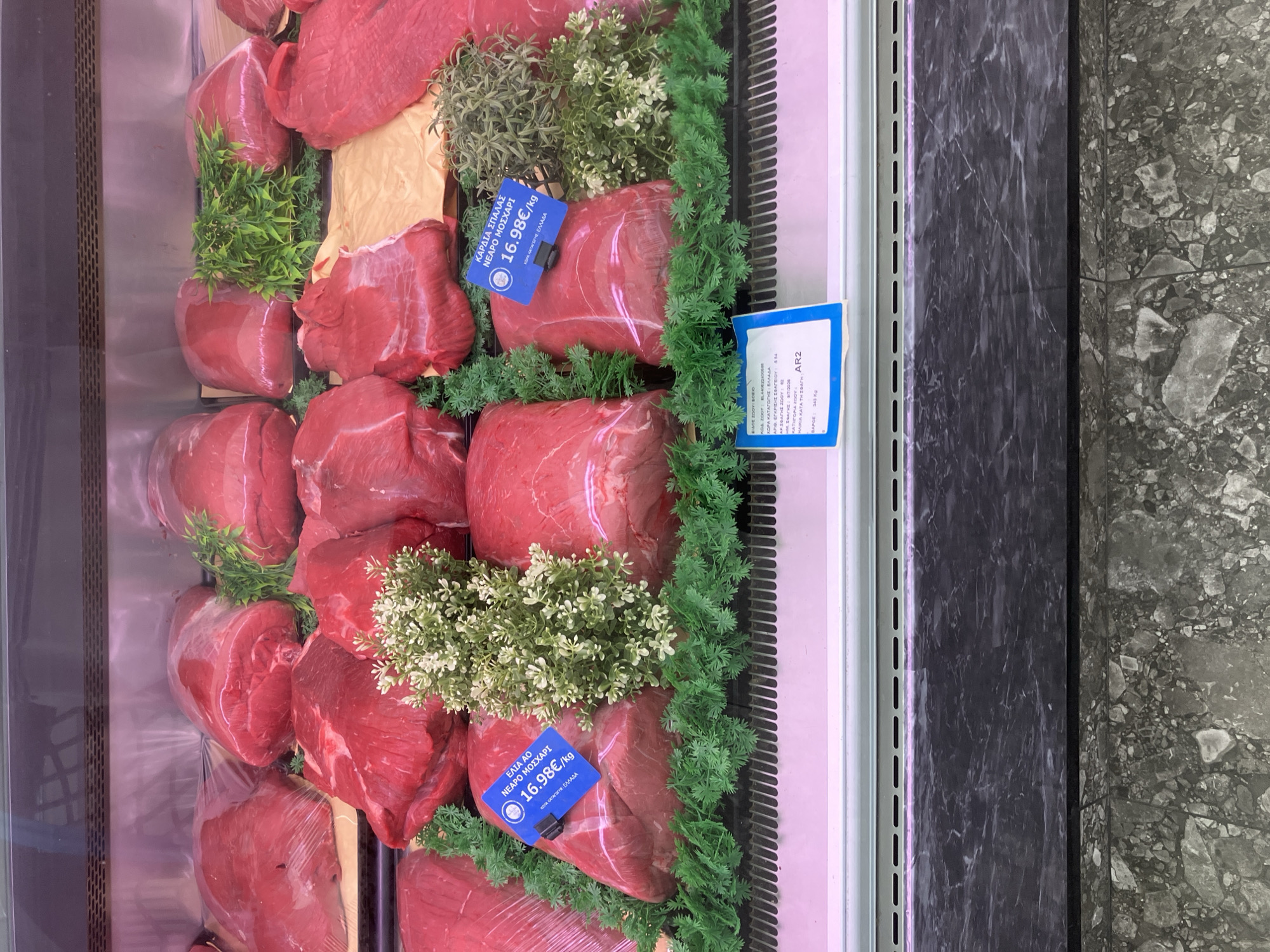 The image shows a display case filled with cuts of raw red meat. The meat is arranged on shelves and is decorated with artificial greenery and small white flowers. There are several blue price tags visible. One tag, on the left side of the image, reads "ΕΔΑ 40 ΝΕΑΡΟ ΜΟΣΧΑΡΙ 16.98€/kg" and "ΧΩΡΑ ΚΑΤΑΓΩΓΗΣ: ΕΛΛΑΔΑ". Another tag, slightly to the right, reads "ΚΑΡΔΙΑ ΣΠΑΛΛΑΣ ΝΕΑΡΟ ΜΟΣΧΑΡΙ 16.98€/kg" and "ΧΩΡΑ ΚΑΤΑΓΩΓΗΣ: ΕΛΛΑΔΑ". The background appears to be the interior of a refrigerated display unit. The bottom of the image shows the exterior of the display case with a marbled surface. The location is specified as Paleo Faliro, Greece, suggesting this is a butcher shop or a meat counter in a supermarket. There are no people visible in the image. The lighting is bright, highlighting the fresh appearance of the meat.