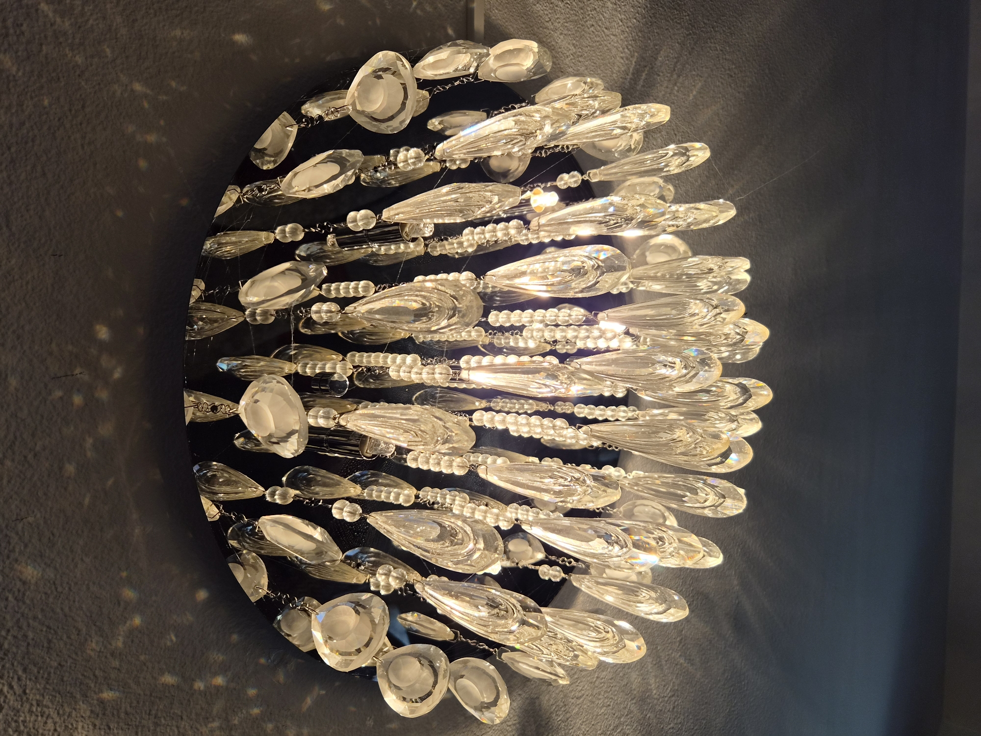 The image displays a close-up, downward-facing view of a decorative chandelier. The chandelier is circular and has a dark base from which numerous strands of clear, teardrop-shaped acrylic or glass crystals hang, interspersed with smaller round beads. These crystals are reflecting the light, creating sparkling highlights and prismatic effects on the dark, textured ceiling above and around the fixture. The overall impression is one of elegance and subtle illumination. There are no people or animals visible in the image. The specific location within Heberg, Sweden, is not discernible from the visual information provided.