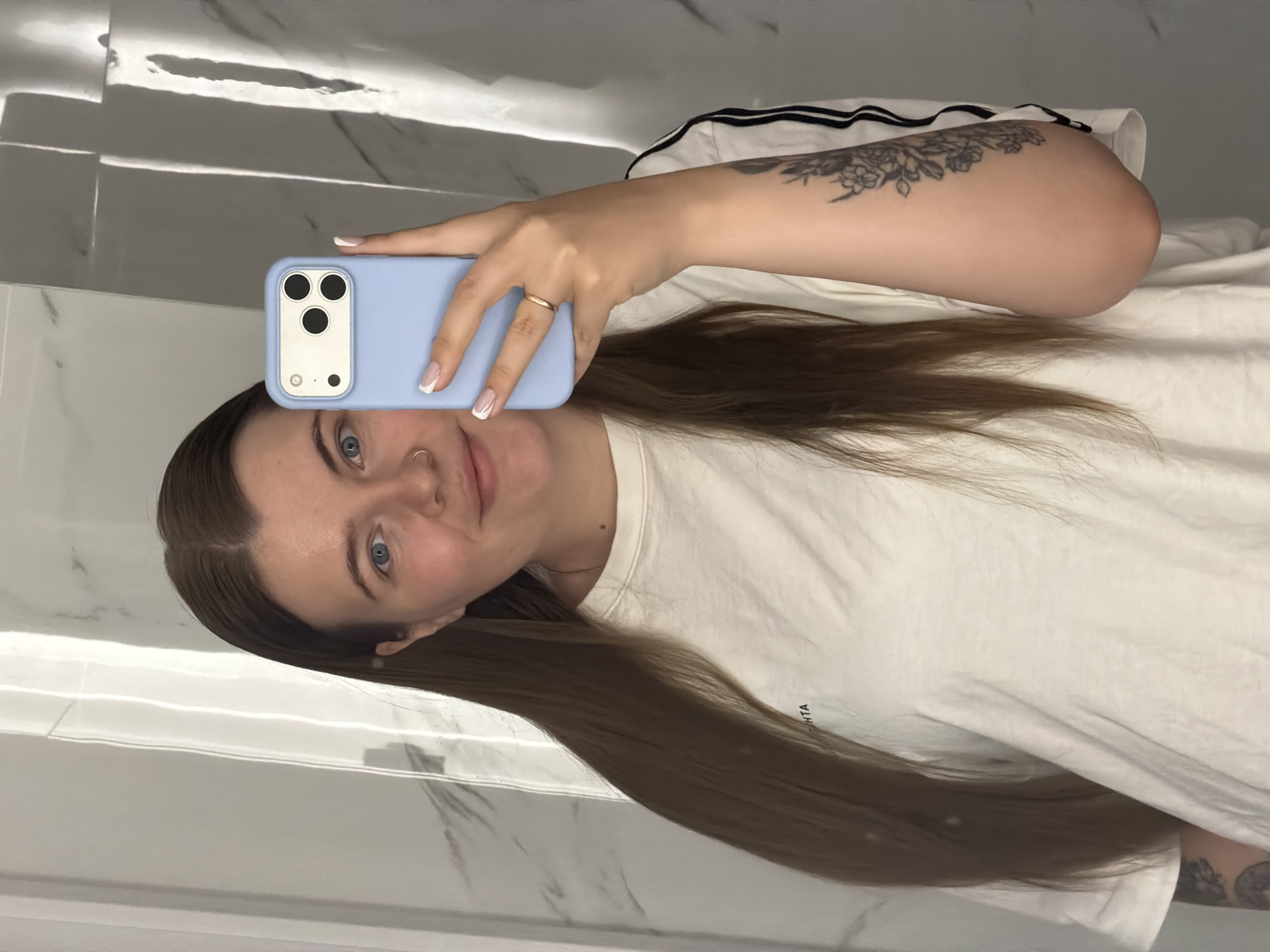 A woman is taking a selfie in what appears to be a bathroom. She is holding a light blue iPhone with a white case over her face. She has blue eyes and brown hair. She's wearing a white t-shirt with a black stripe on the sleeve and a tattoo on her left arm. A gold ring is on her finger. The background is a white marble wall. This image does not provide information about location in Chaikovsky, Russia.