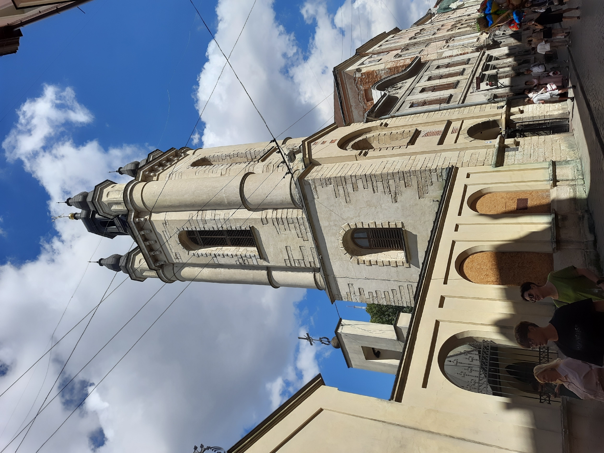 The image captures a view of the Armenian Cathedral in Lviv, Ukraine, on a sunny day with scattered clouds. The cathedral's tall, light-colored bell tower dominates the center of the frame, featuring multiple levels and arched windows. Adjacent to the tower is a smaller bell structure topped with a cross. To the left, a light yellow wall with arched openings, one featuring a decorative metal gate, runs along the street. Some of these arched openings are boarded up.
In the foreground, a paved street is visible with a few people walking. On the left, two women are walking towards the viewer, one with blonde hair wearing a pink blouse, and the other with dark hair wearing a green top and sunglasses. In the middle, a person wearing glasses and a black t-shirt is walking. Further down the street, on the right, a group of people are gathered, and some are walking away from the viewer. A person is holding a bunch of colorful balloons. The buildings lining the street exhibit varied architectural styles, with some showing signs of age and repair, particularly the one to the right of the bell tower with exposed brickwork. Overhead, several electrical wires crisscross the sky. The scene depicts a typical urban streetscape with historical architecture and pedestrian activity.