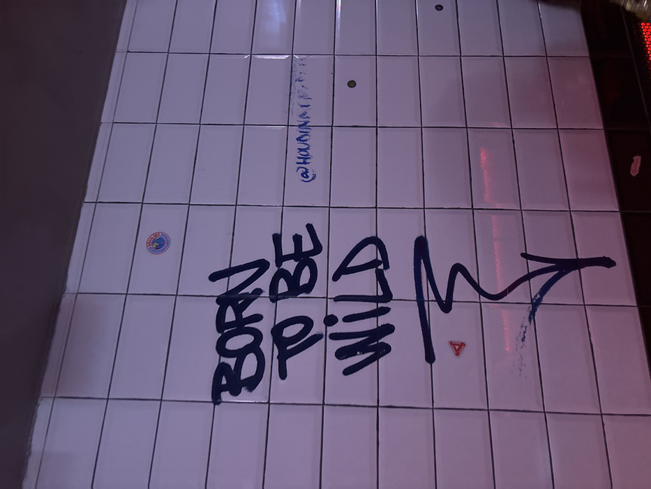 The scene depicts a white tiled wall in a Madrid Metro station. The wall is constructed from rectangular white ceramic tiles with dark grout lines. Prominently spray-painted in thick, dark blue or black letters on the center-left is the phrase "BORN TO BE WILD," with a stylized downward-pointing arrow or squiggle beneath it. To the right of this, in a lighter blue, is a smaller, partially legible tag beginning with "@HOUDINI_". A circular "Metro de Madrid" logo sticker, featuring a red and blue design with a white 'M', is affixed to a tile in the upper central area. A small, red triangular sticker is located below "WILD". Two brass-colored circular recessed fittings or holes are visible on the right side of the wall. The environment is illuminated by artificial light, casting a distinct purple and pink ambient hue across the tiles, especially noticeable towards the bottom where a horizontal black trim element runs along the wall's base. No individuals, actions, or interactions are present in the image.