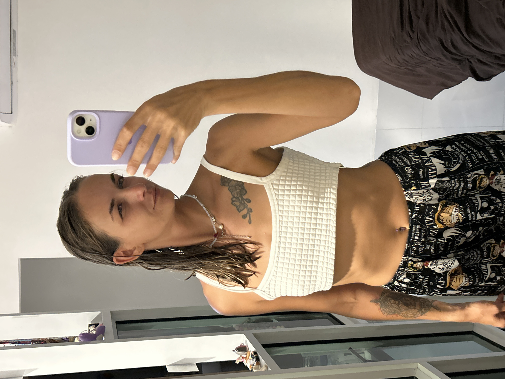 In a room in Kathu, Thailand, a woman with wet hair takes a mirror selfie. She is wearing a white waffle-knit crop top, patterned pajama bottoms, and a delicate necklace. She is holding a lavender phone in her right hand. Tattoos are visible on her chest and left arm. The background shows a white wall, a window with shelves, and a dark brown surface that appears to be a bed.