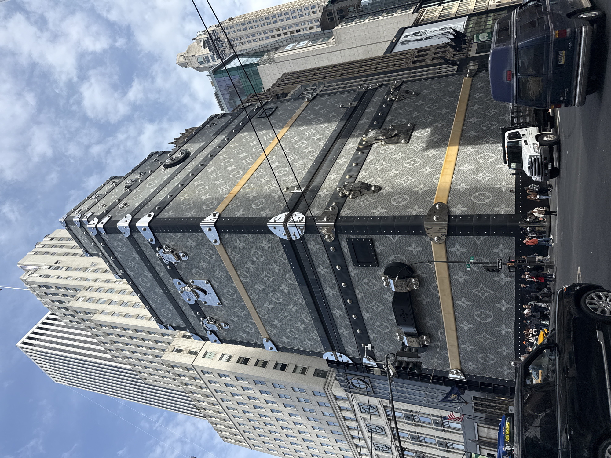 The image depicts a large Louis Vuitton trunk placed on a city street in New York City, United States of America. The trunk's exterior is covered in the brand's signature monogram pattern. Several people are standing near the trunk, along with parked cars and vans. Buildings typical of Manhattan's architecture rise in the background, and overhead power lines are visible. The sky above is partially cloudy.