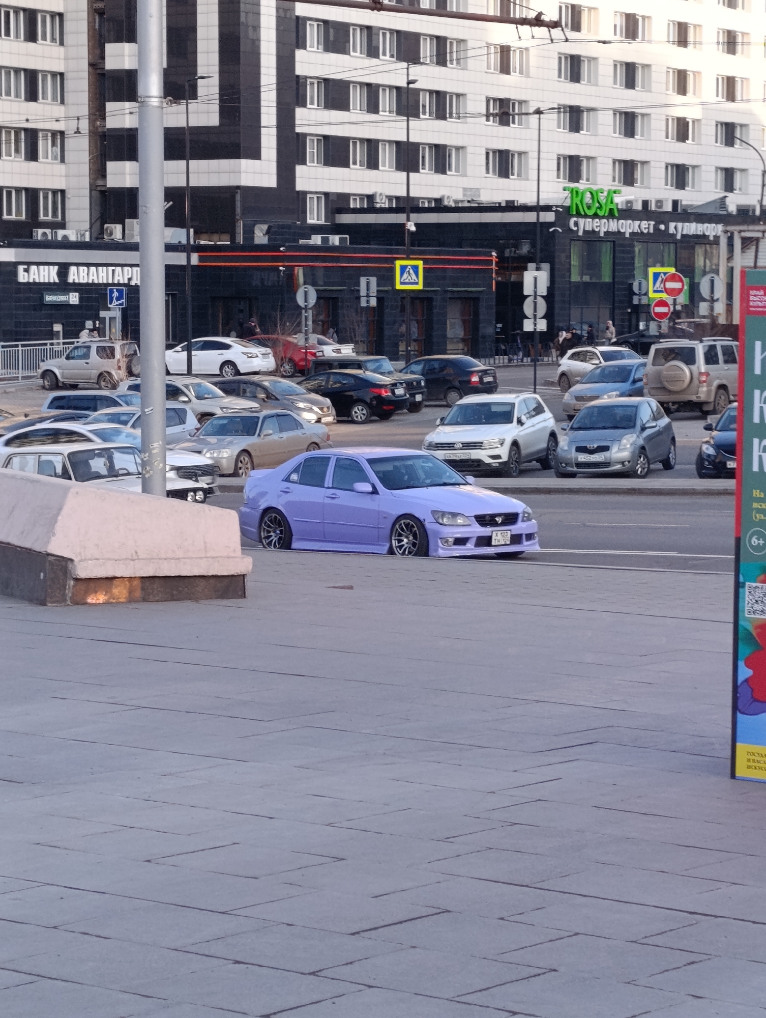 The image shows a lilac sedan on a city street in Krasnoyarsk, Russia. The car is heavily modified and has aftermarket wheels. Other cars are parked and driving nearby. "БАНК АВАНГАРД" and "ROSA супермаркет - кулинар" signage are visible on buildings in the background. Pedestrians are present near the businesses. The day appears to be overcast.