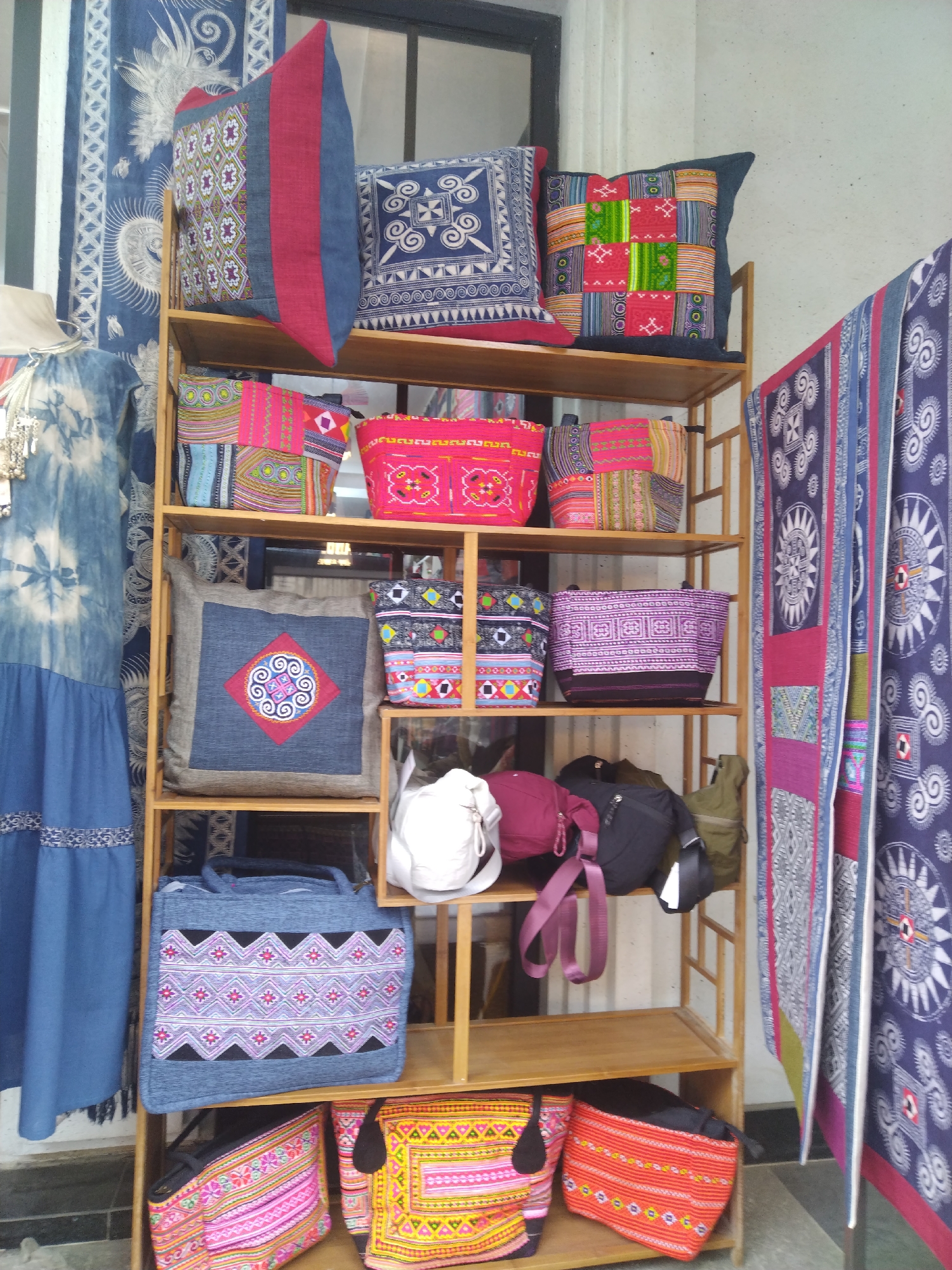 This image captures a vibrant display of traditional textile products, likely for sale, within a market stall or shop in Sa Pa Ward, Vietnam. The scene is dominated by a tall, wooden shelving unit filled with a variety of handcrafted items, flanked by other textile goods.
The main subjects are numerous pillows and bags, characterized by intricate patterns and a rich palette of colors including red, blue, green, pink, purple, white, yellow, orange, and black. Many items feature geometric and folk-art inspired designs, some incorporating patchwork techniques and denim-like fabric accents. Several pillows are arranged on the top shelves, displaying bold patterns. Below, various tote bags, shoulder bags, and smaller pouches are neatly stacked or hung, also showcasing diverse colorful patterns.
To the left of the shelves, a blue dress with a tie-dye top and a solid skirt with a patterned hem hangs on a partially visible mannequin. To the right, multiple fabric hangings or tapestries, predominantly in blue/purple with white and red patterns, are displayed vertically. The wooden shelves themselves are simple and made of light-colored wood. In the background, a light-colored wall and what appears to be a window are visible.
The activity taking place is the retail display of traditional handicrafts. The distinct patterns, vibrant colors, and patchwork styles are characteristic of the textile traditions of ethnic minority groups in the northern regions of Vietnam, strongly indicating the specified location context of Sa Pa. The lighting suggests it is daytime. There is no visible text legible within the image. The overall impression is one of cultural richness and skilled craftsmanship.