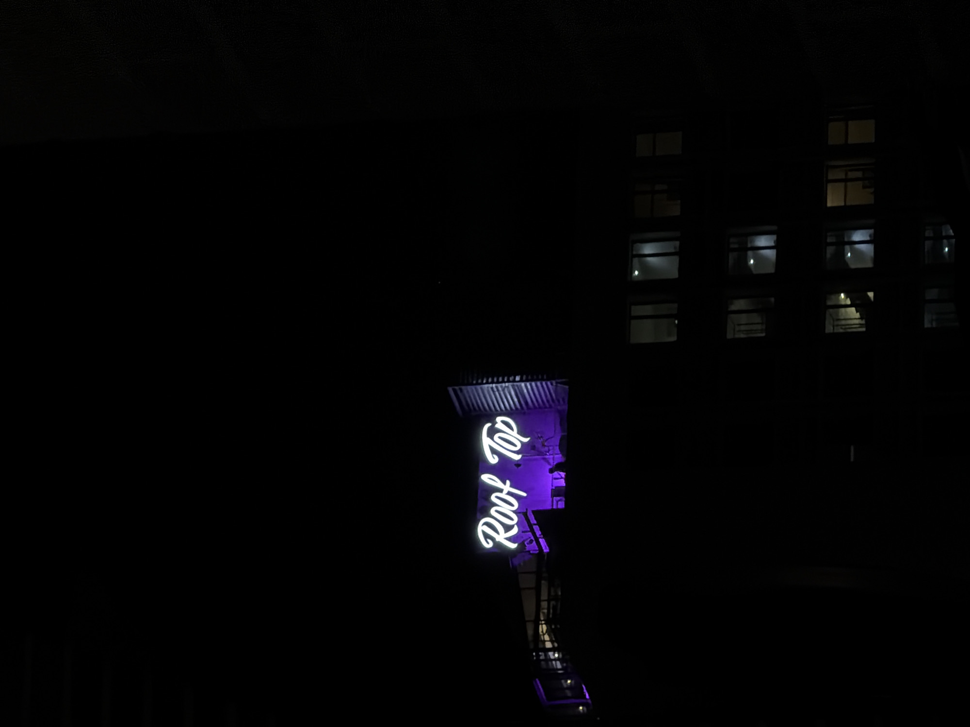 At night, the illuminated neon sign "Rooftop" is visible in Gdańsk, Poland. The sign is vertically oriented on a structure and emits a bright purple/white light. In the background, a building with illuminated windows can be seen, though the overall scene is quite dark. The photograph is taken from a low vantage point.