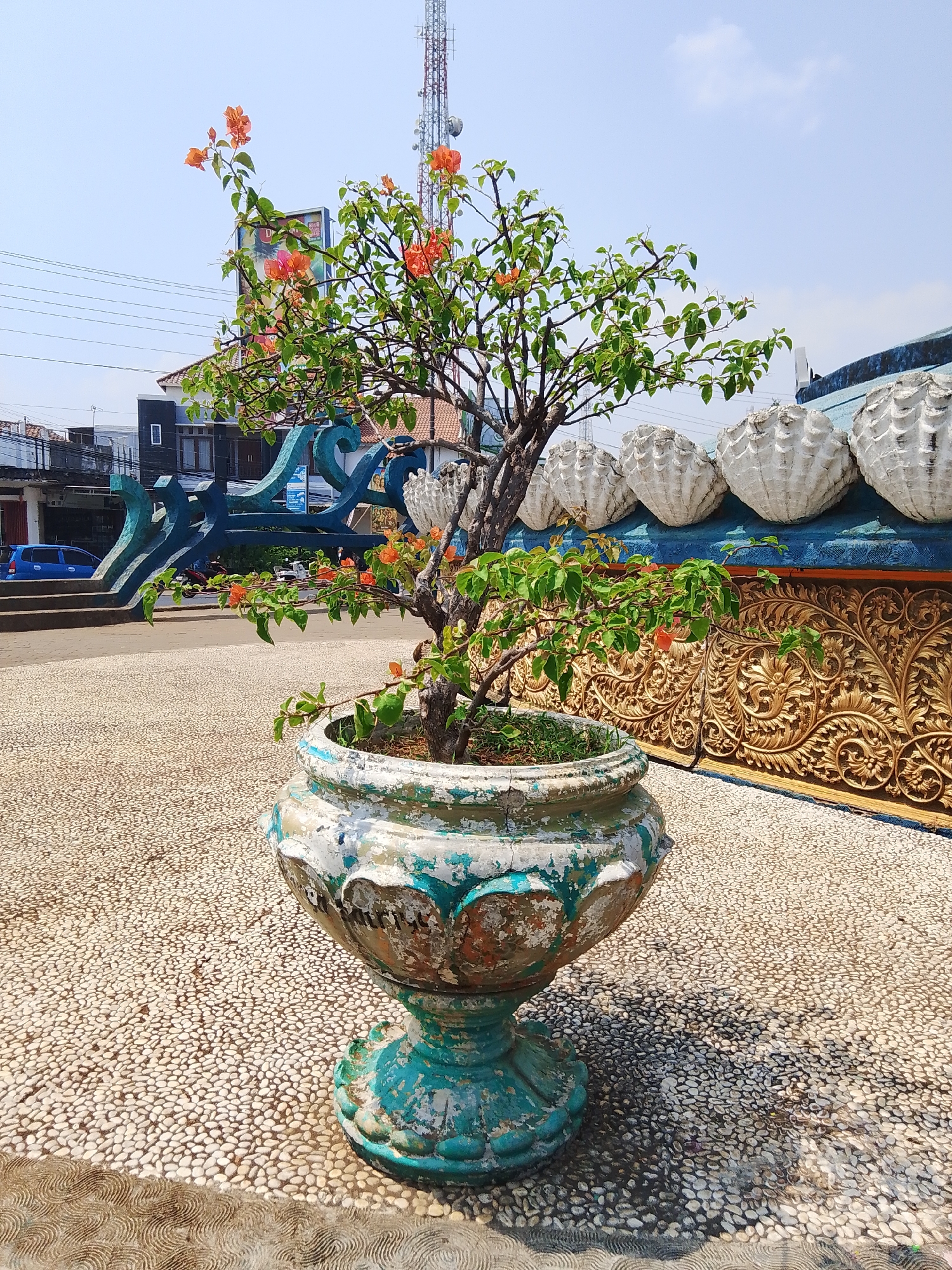 In Tahunan, Indonesia, a potted bougainvillea with vibrant orange flowers stands prominently on a pebble-covered ground. The pot itself is weathered and ornate, painted in shades of turquoise and white, showing signs of age and wear. Behind the plant, a decorative wall features a series of white, shell-like moldings and an intricate golden relief pattern. The background reveals a street scene with buildings, power lines, a telecommunications tower, and a blue car. The sky above is clear and bright blue with a few wisps of clouds.