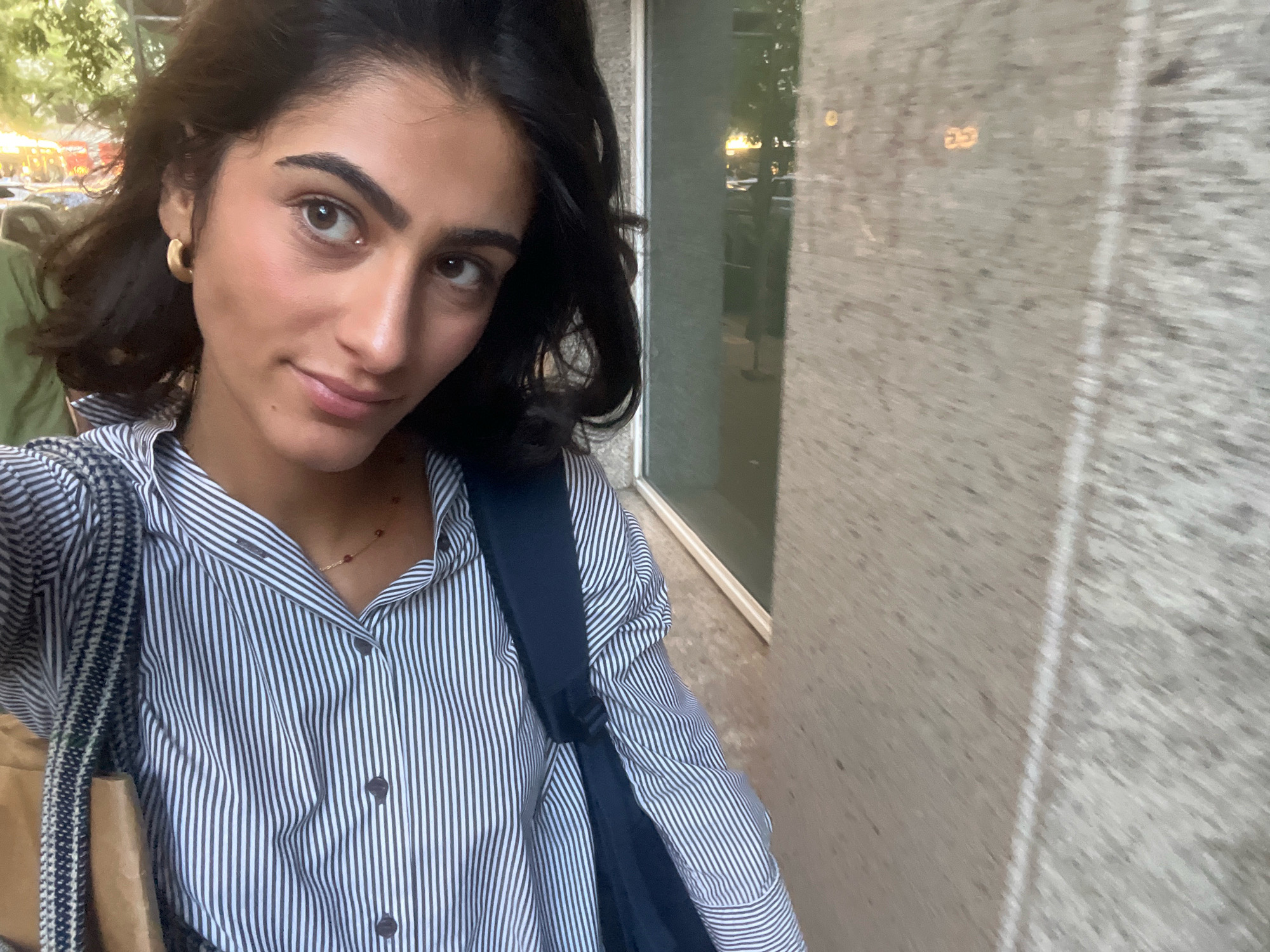 A young woman with dark hair is taking a selfie. She is wearing a black and white striped button-down shirt and a gold hoop earring. She has a dark backpack on her shoulder. She is standing in front of a building with a textured grey facade and a large window. Reflected in the window are trees and streetlights. The image appears to have been taken during the day. The overall scene is typical of an urban environment. The woman's pose and direct gaze suggest she is aware of being photographed. The presence of a backpack indicates she may be on her way to or from a location. The background of trees and streetlights suggests an outdoor setting within a city. The woman's attire is casual yet neat.