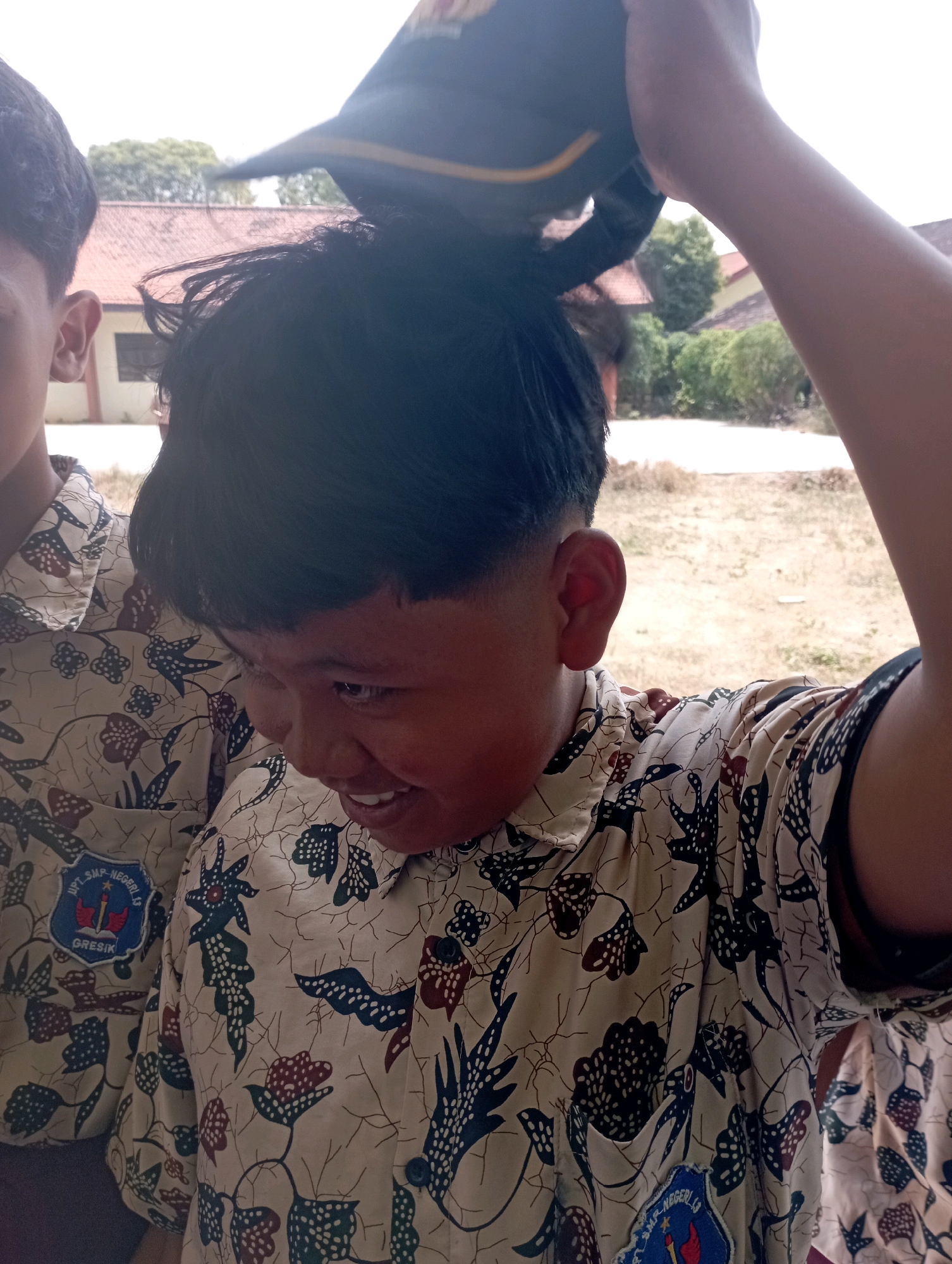 In Banyuurip, Indonesia, two young boys are pictured outdoors. The boy in the foreground, wearing a tan batik shirt with a dark blue and red emblem on the left chest pocket, is looking down and to the left, smiling. He has short, dark hair and is holding a dark blue and yellow cap above his head with his right hand. Another boy, partially visible on the left, also wears a similar batik shirt. The background suggests a dry, outdoor environment with sparse vegetation and buildings with terracotta roofs.