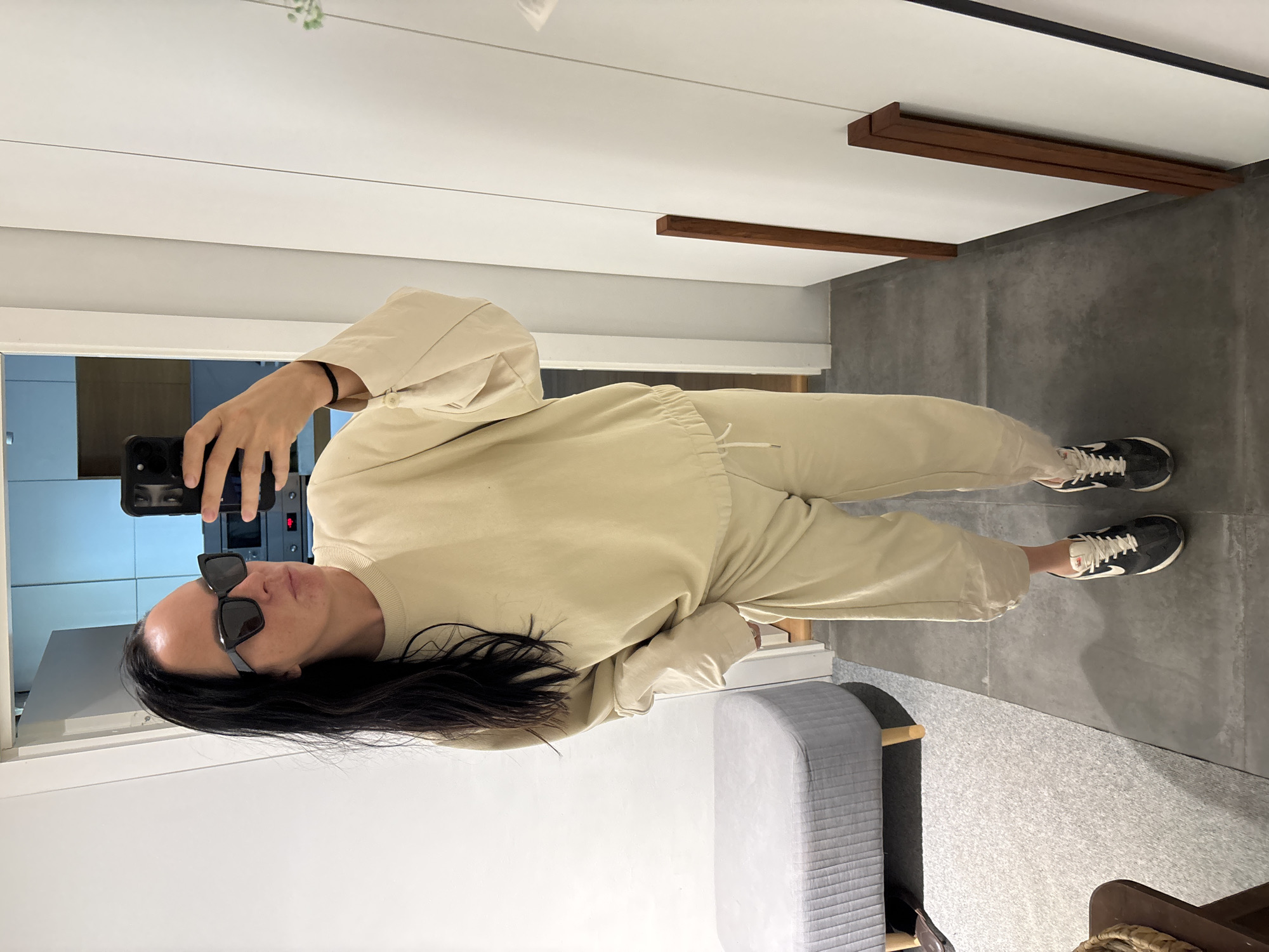 A person wearing sunglasses, a beige matching sweat suit, and black and white sneakers is taking a selfie in front of a mirror. The person is holding a black smartphone. Behind the person, there is a room with a light gray bed and a light gray concrete floor. A glimpse of a kitchen with light blue cabinets can be seen in the mirror's reflection. There is no information in the image to indicate that this photo was taken in the city of Chaikovsky, Russia.