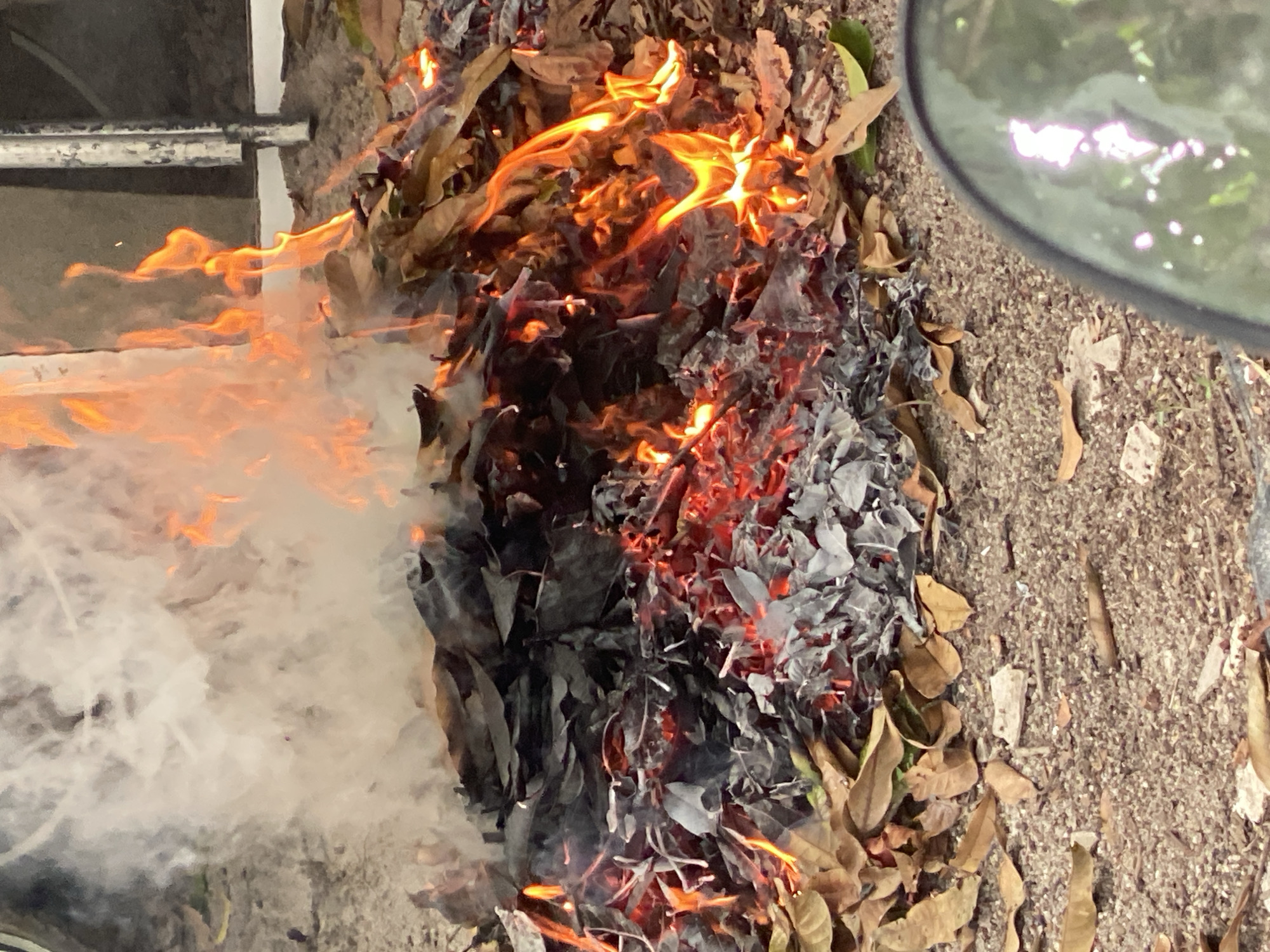 A pile of burning leaves is located on the ground. Flames rise from the center of the pile, and white smoke is visible to the left. The leaves range in color from brown to black and grey. The ground is a mixture of dirt and small rocks. A partially visible metal structure is in the upper left, and a shiny, round object is in the upper right.