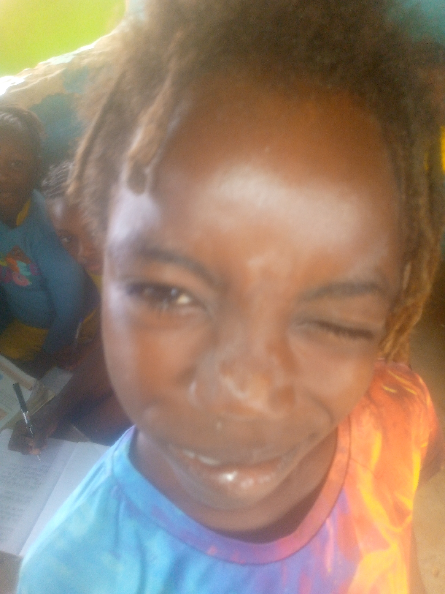 A young, dark-skinned person with light brown dreadlocks winks at the camera. They wear a tie-dye shirt with blue, purple, orange, and yellow coloring. The background is blurred, showing two other children and a pen in hand, suggesting a classroom setting. The location is specified as Unknown, Unknown.