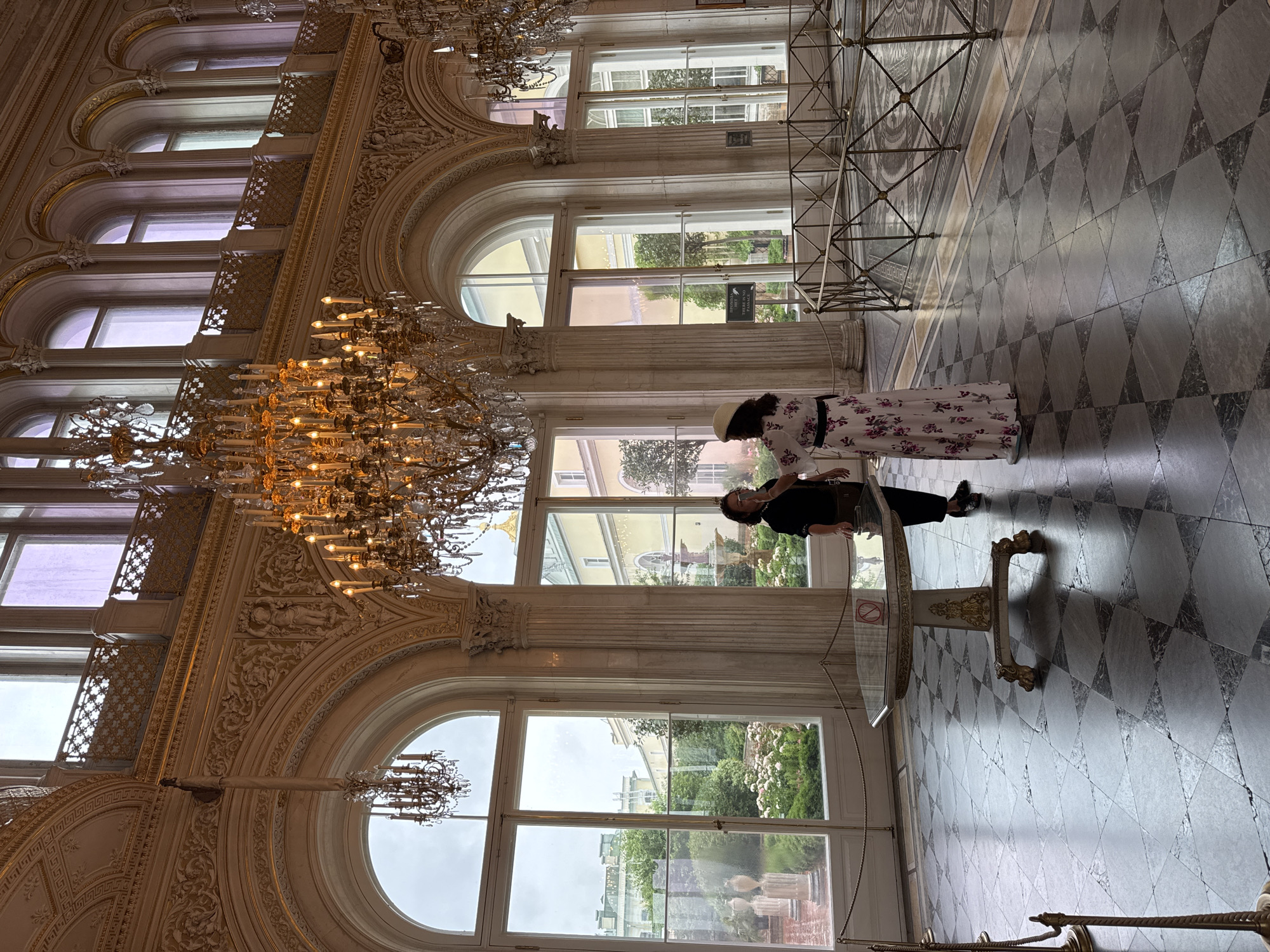 The image shows two people in a grand, opulent hall with high ceilings and large windows. The hall features ornate architectural details, including intricate carvings and large chandeliers. The floor is tiled with a diamond pattern of light and dark grey marble.
One person, a woman, is wearing a long floral dress and a hat. She is standing near a large round glass table with an ornate base. The second person, a man, is seated at the table, leaning forward, and appears to be interacting with a tablet or phone. The scene is indoors, and the large windows offer a view of lush green gardens and buildings outside, suggesting it is daytime. The lighting in the hall is a mix of natural light from the windows and the warm glow from the chandeliers, creating a somewhat formal and elegant atmosphere.
The location is identified as Saint Petersburg, Russia. There are some visible signs on the windows, but the text is not clearly legible. The overall impression is one of visiting a historic and well-preserved grand interior.