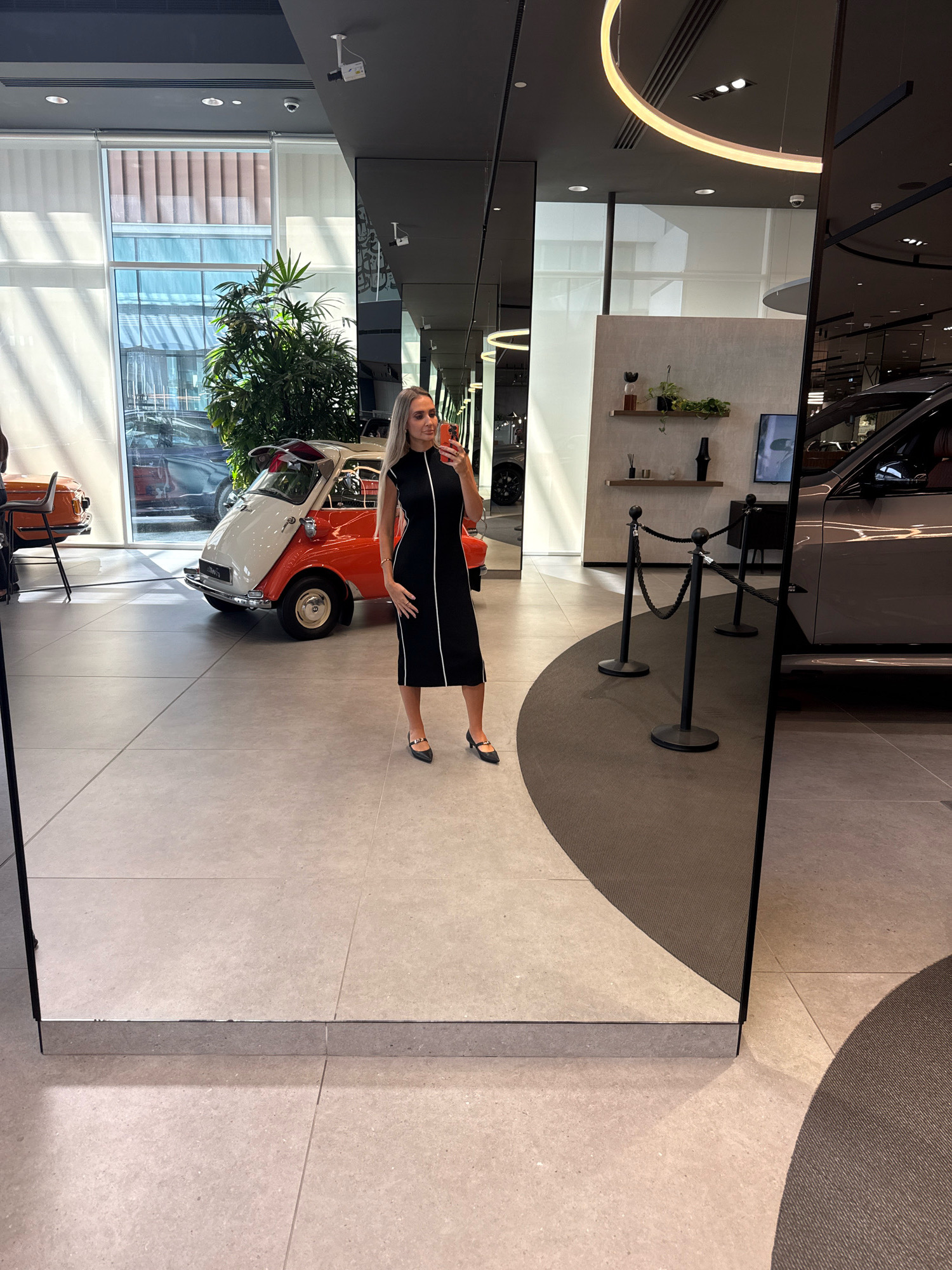 A woman with blonde hair is taking a mirror selfie in what appears to be a modern car showroom. She is wearing a black, sleeveless, knee-length dress with white trim. She is holding an orange smartphone in her right hand and standing in front of a large mirror.
In the reflection, to her left, is a vintage two-toned orange and white microcar. Behind the microcar, large windows reveal a city skyline with tall buildings, suggesting the location is in Dubai, United Arab Emirates. To her right in the reflection is a modern grey SUV. The showroom floor is tiled with a light-colored material, and a dark grey carpeted area with stanchions is visible in the foreground. The ceiling features sleek lighting fixtures. The overall environment is clean, contemporary, and well-lit.