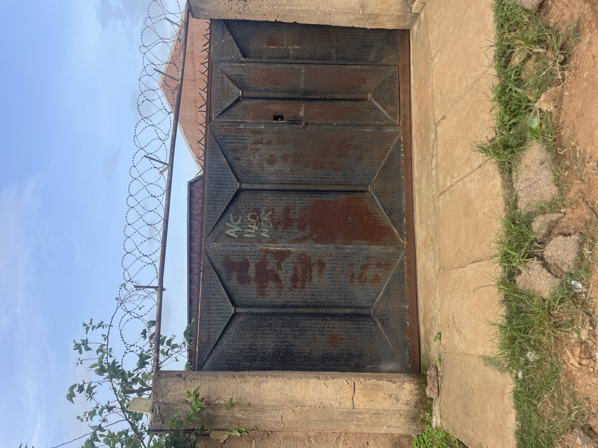 The image shows a closed, double metal gate in Bukuru, Nigeria. The gate is dark gray with a diamond plate texture and appears to be rusted in places. There is some writing in green spray paint on the right panel of the gate. The gate is flanked by two concrete pillars, the left one of which has some green vines growing on it. Above the gate, a line of barbed wire is stretched between two metal posts. In the background, a red corrugated metal roof is visible. The ground in front of the gate is a concrete walkway with some patches of grass and dirt. The sky is cloudy.
