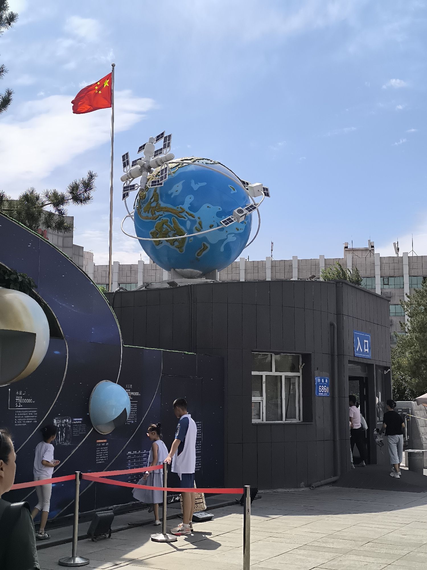 The image depicts an outdoor scene at the Xinjiang Astronomical Observatory in Ürümqi, China. In the foreground, a large, blue globe representing Earth, adorned with a satellite model, is displayed prominently. A Chinese flag flies on a flagpole to the left of the globe. A group of people, including children and adults, are observing an exhibit on the wall behind a red rope barrier. The exhibit features diagrams and information about planets and space. To the right, an entrance to a building is visible, with people entering and exiting. The architecture suggests a modern facility, likely part of the observatory. The sky is clear blue with scattered clouds, indicating a sunny day. The overall impression is of an educational and public-facing space, likely a visitor center or museum associated with the astronomical observatory.