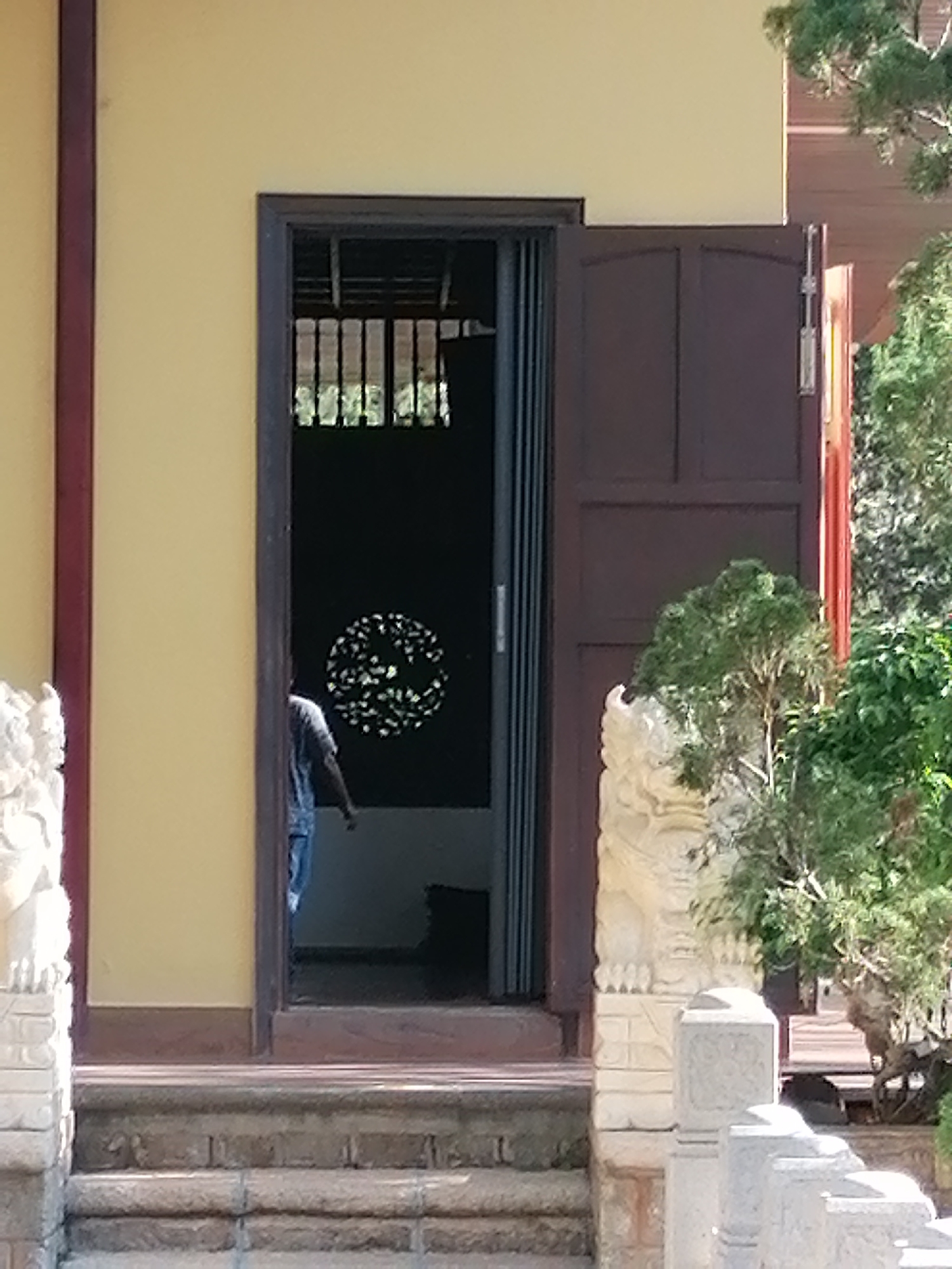 The image shows the entrance to a building with a yellow facade and a dark brown double door, one of which is ajar. A person in a blue t-shirt and jeans is walking into the building. Inside, there is a circular decorative element on the wall. To the left of the entrance, there is a stone statue and stairs leading up to the building. To the right, there is a small tree and more stone sculptures. The setting appears to be a temple or similar cultural site. This scene is located at the Truc Lam Zen Monastery in Da Lat, Vietnam, a prominent tourist attraction known for its serene atmosphere and beautiful architecture.