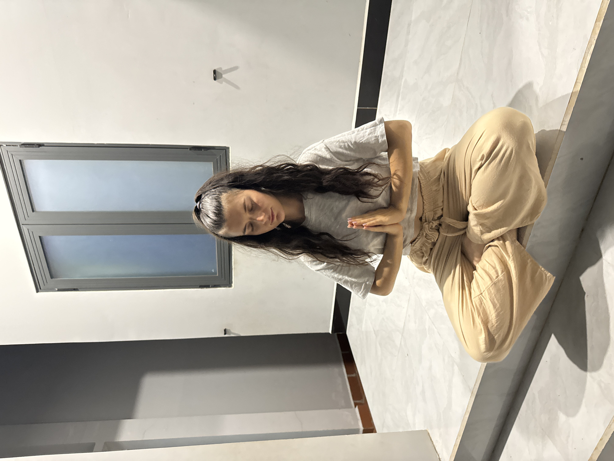 A young woman with long dark hair is seated in a meditative pose with her eyes closed and hands pressed together. She is wearing a light gray t-shirt and light tan pants. The setting appears to be an indoor space with a modern architectural style, including a window and a gray wall detail. The scene is lit with ambient lighting. The location is Mũi Né, Vietnam.