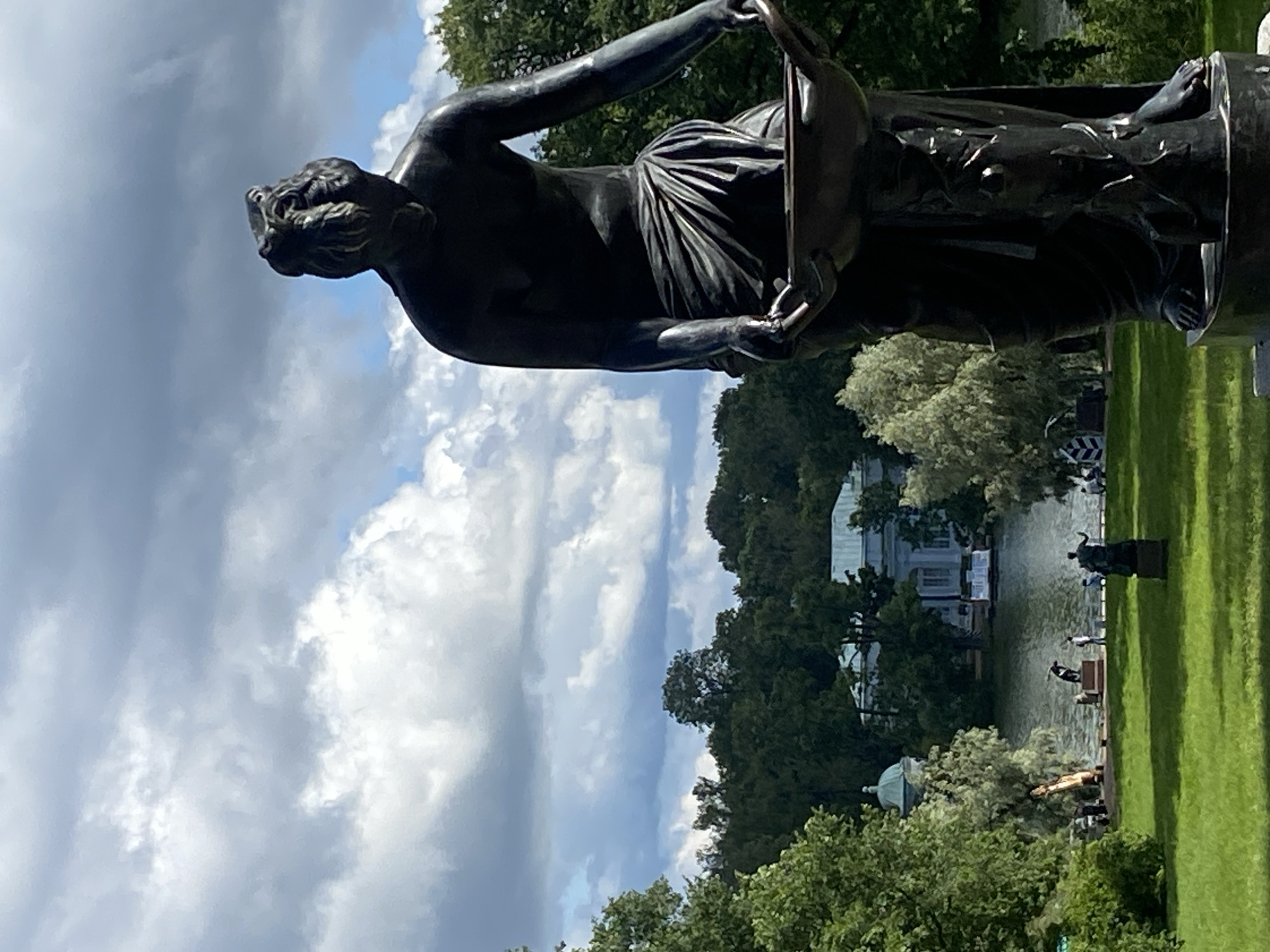 This is a full-frame, eye-level, vertical shot of a dark bronze statue of a draped figure holding a shallow bowl. The statue is in the foreground, on the right side of the image, and is the primary subject. Its face is obscured, and it appears to be wearing a toga-like garment. Behind the statue, on the left, is a bright green lawn and further back, a body of water, likely a pond or lake, surrounded by dense green trees. In the distance, across the water, is a light-colored neoclassical building with a green dome and a pale blue facade. The sky is overcast with dramatic, dark grey clouds interspersed with patches of white. Several other smaller statues and a few people are visible on the far side of the lawn, near the water's edge. One person is standing near a larger, lighter-colored statue on the right side of the far lawn, and another group of people is near the water. The overall setting appears to be a park or garden. The architectural style of the building and the presence of statues suggest this scene is located in Pavlovsk, Russia, likely within the Pavlovsk Park, a historic landscape park that is part of the Pavlovsk Palace ensemble.