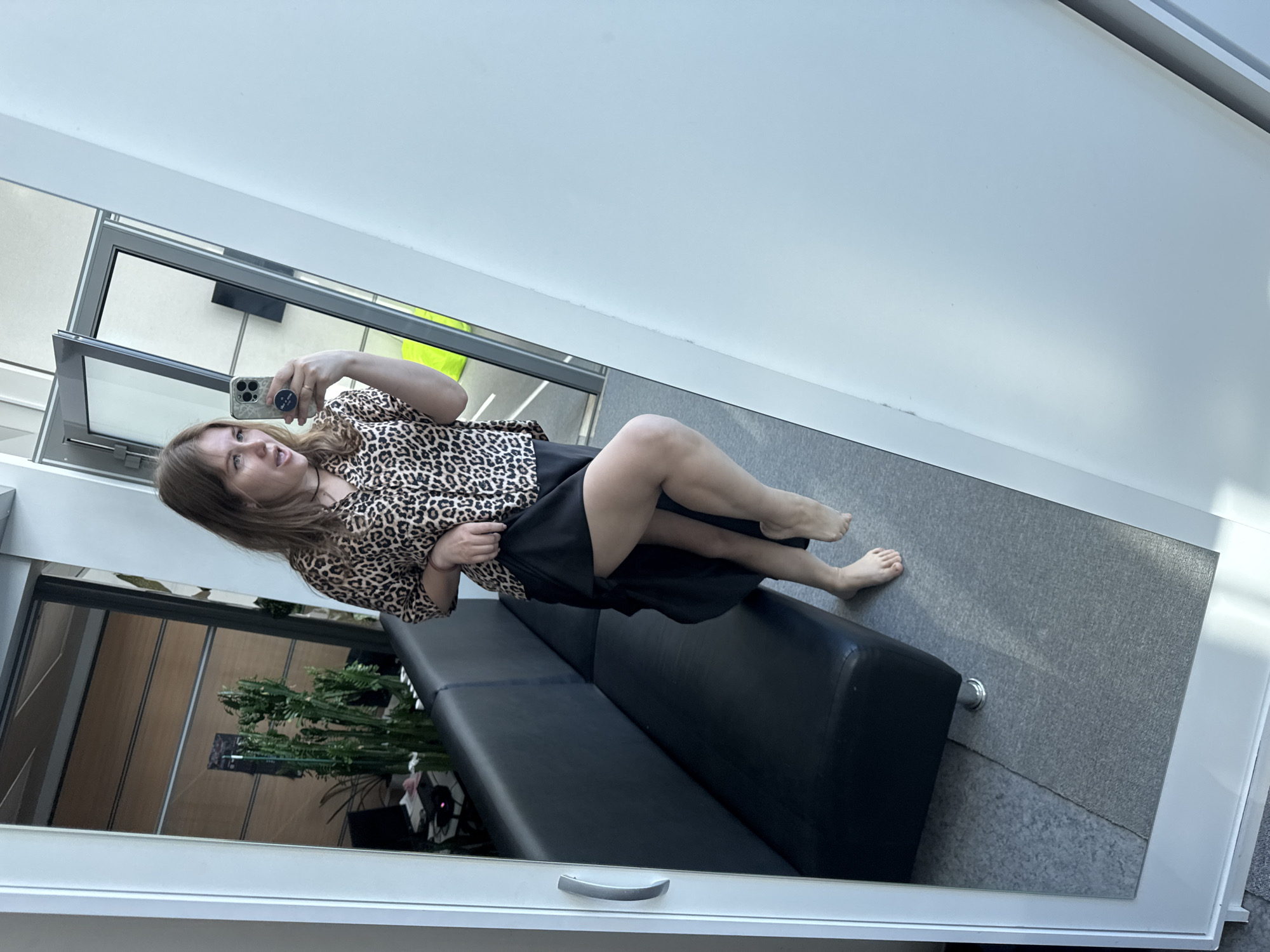 A young woman is taking a mirror selfie. She is wearing a leopard print top and a black skirt. She is sitting on a black leather couch. In the reflection of the mirror, a window reveals a potted plant. The floor is carpeted in gray. The location could be Perm, Russia, but this cannot be definitively determined from the image.