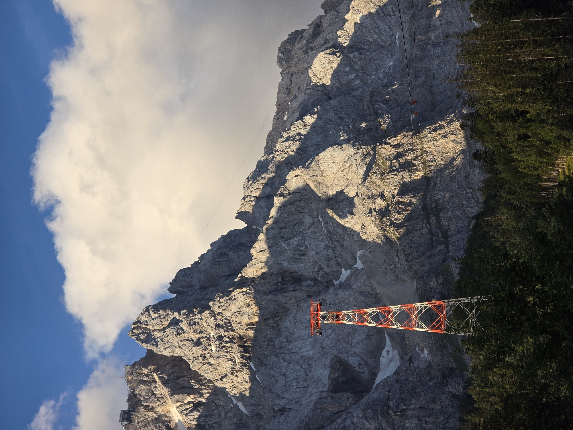 The image captures a mountainous landscape in Grainau, Germany, specifically depicting a section of the Zugspitze massif. In the foreground, a dense forest of evergreen trees covers the lower slopes. Rising above the trees is a prominent red and white lattice tower, likely a support structure for a cable car system. In the background, the rugged, rocky face of the mountain dominates the scene. The peak is partially obscured by large, white clouds, with patches of blue sky visible above. The sunlight casts dramatic shadows across the mountainside, highlighting its textured, uneven surface. Faint lines of a cable car can be seen stretching across the mountain, disappearing into the clouds. There are no people or animals visible in the frame. The scene conveys a sense of grandeur and the imposing scale of the natural environment.