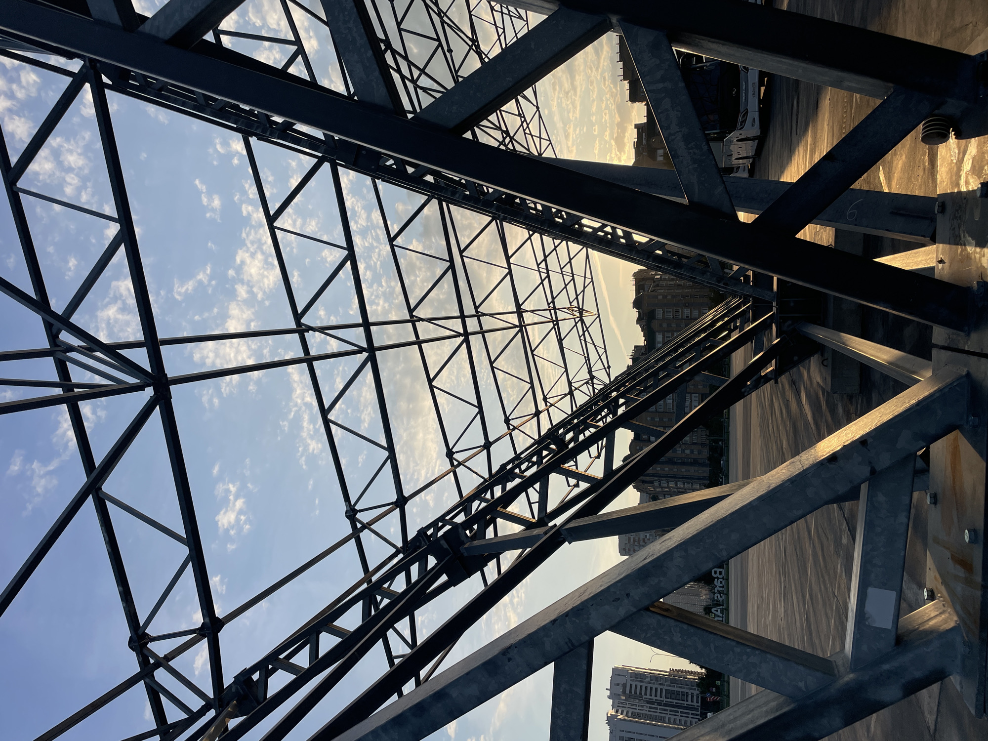 This is a photograph of a metal truss structure, likely part of a covered parking area or bus station, against a bright sky with scattered clouds. The sun is low in the sky, casting long shadows and golden light on the ground and some of the metal beams. In the background, residential buildings and a white bus are visible. The scene appears to be an outdoor urban environment, possibly in Kazan, Russia, given the signage in Cyrillic alphabet on the ground. There are no people clearly visible in the image. The overall impression is one of industrial architecture meeting a soft, late-afternoon sky.
